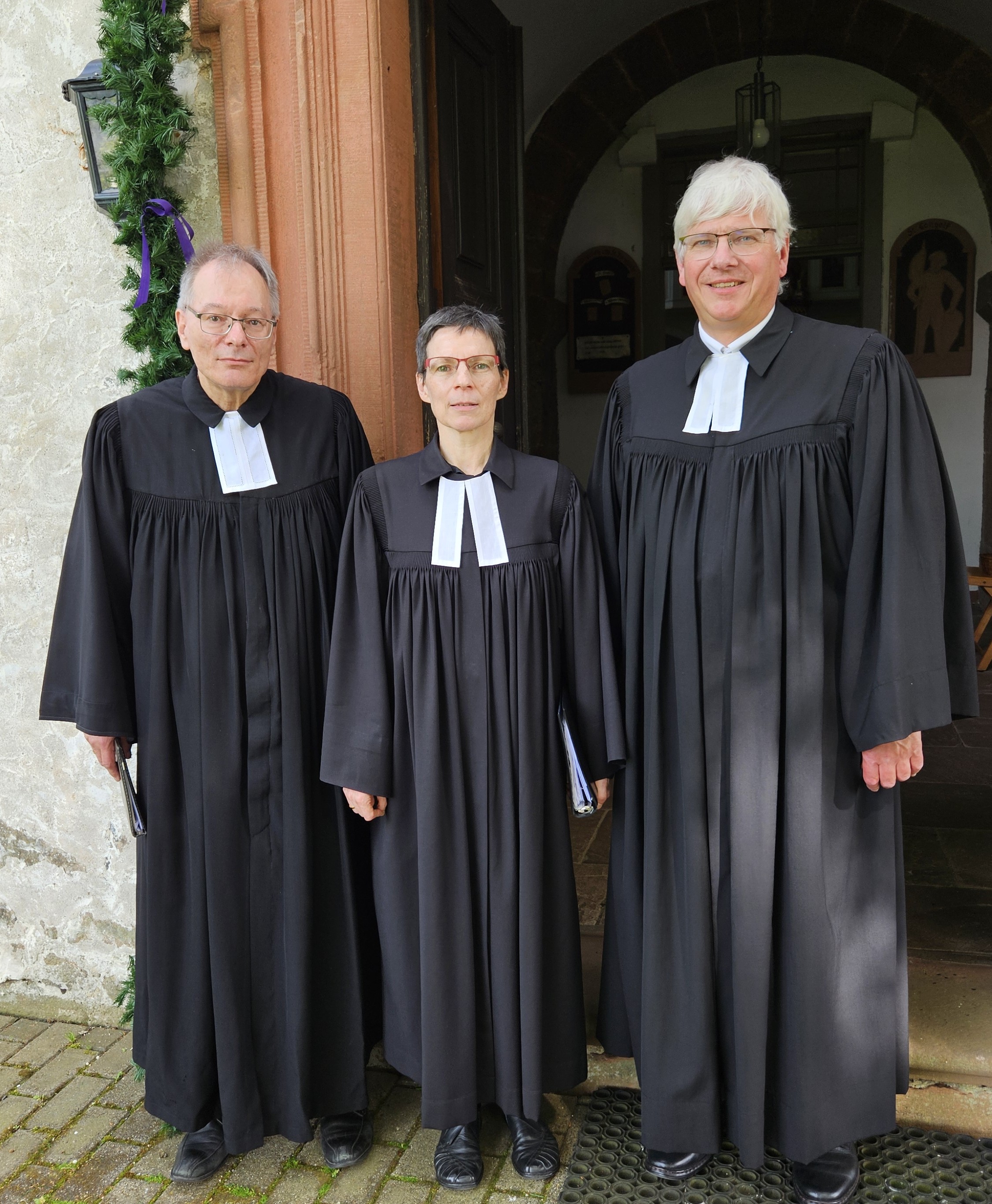 Pfarrer Detlef Metz, Pfarrerin Eleonore Merkel und stellvertretender Dekan Ulrich Bauersfeld stehen vor der geöffneten Tür der Kirche St. Gangolf in Bobenhausen II. Alle drei tragen einen Talar mit Beffchen. Neben der Tür sieht man eine grüne Girlande, die den Eingang schmückt.