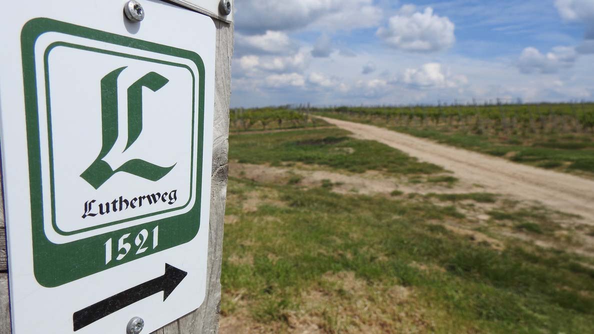 Wegweiser mit der Aufschrift „Lutherweg 1521“ und Pfeil nach rechts; daneben führt ein unbefestigter Weg durch eine offene Landschaft unter bewölktem Himmel.