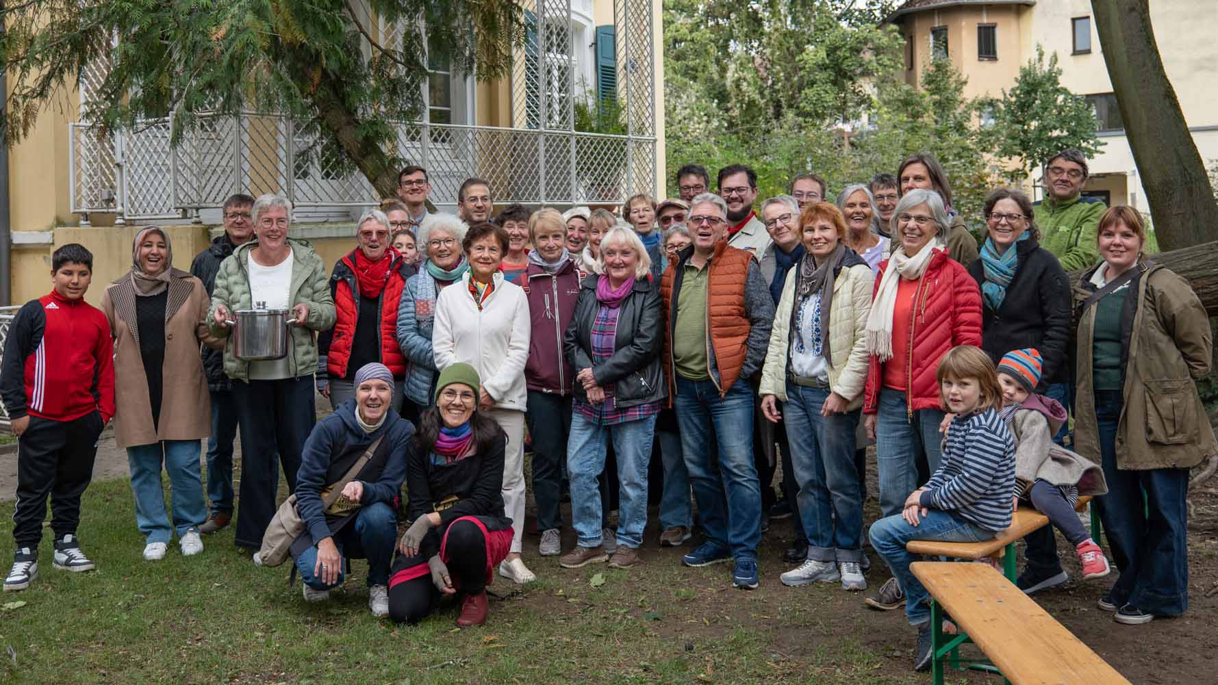 Einheitssuppe_2025_C_Fitz_van_Thom_16zu9.jpg Große Gruppe von Erwachsenen und Kindern steht und sitzt gemeinsam im Garten; eine Person hält einen großen Kochtopf.