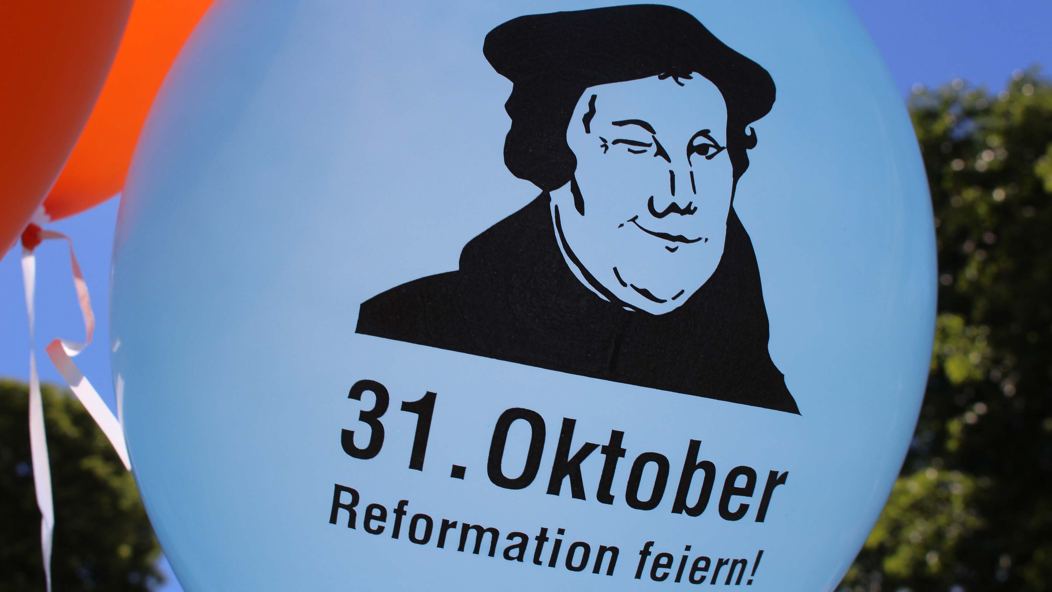 Alternativtext: Blauer Luftballon mit schwarzer Zeichnung von Martin Luther und der Aufschrift „31. Oktober – Reformation feiern!“. Im Hintergrund orangefarbene Luftballons.
