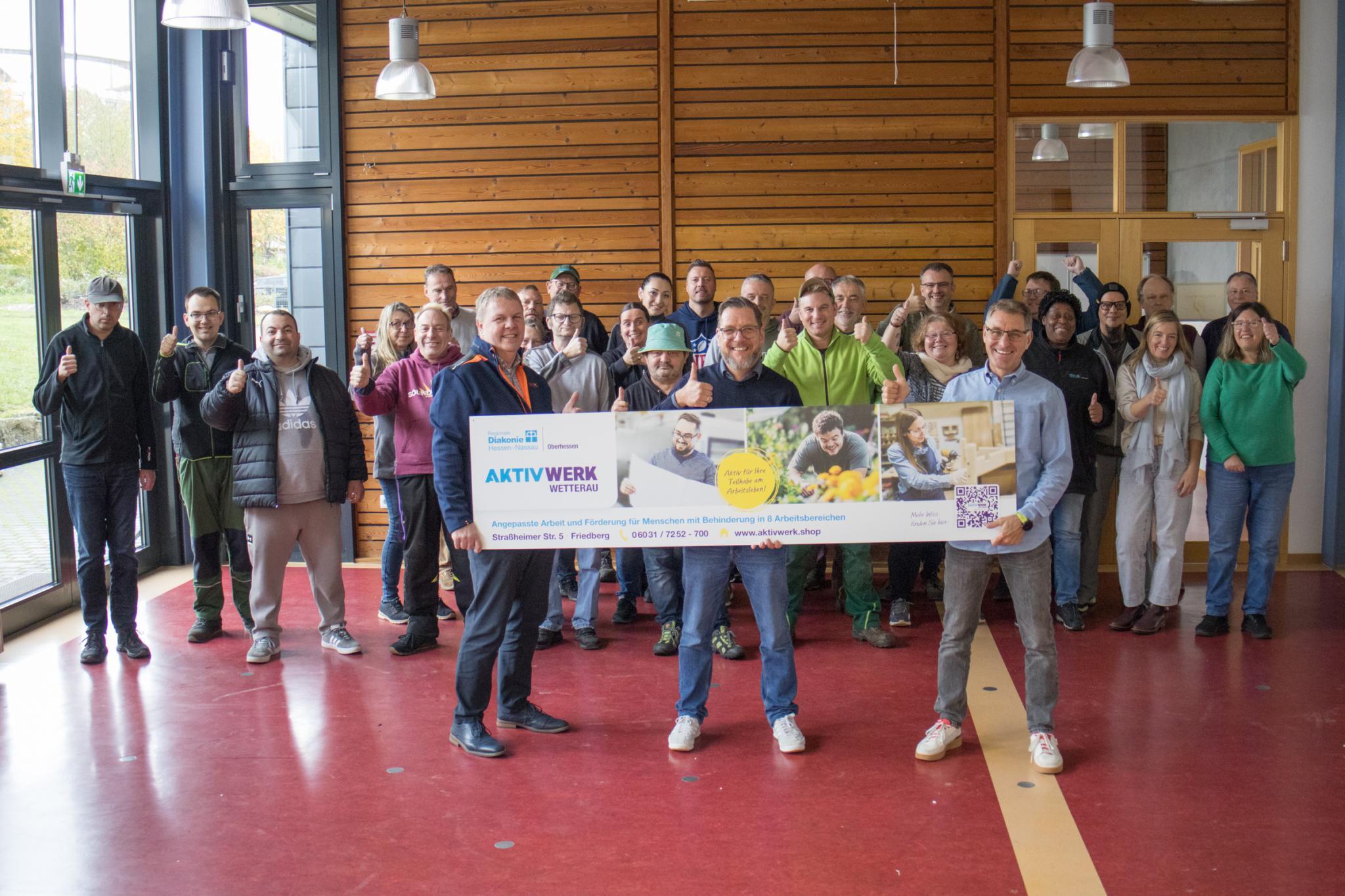 Eine große Gruppe von Menschen steht in einem Raum, drei Männer halten ein großes Transparent der Regionalen Diakonie, alle anderen recken den rechten Daumen,