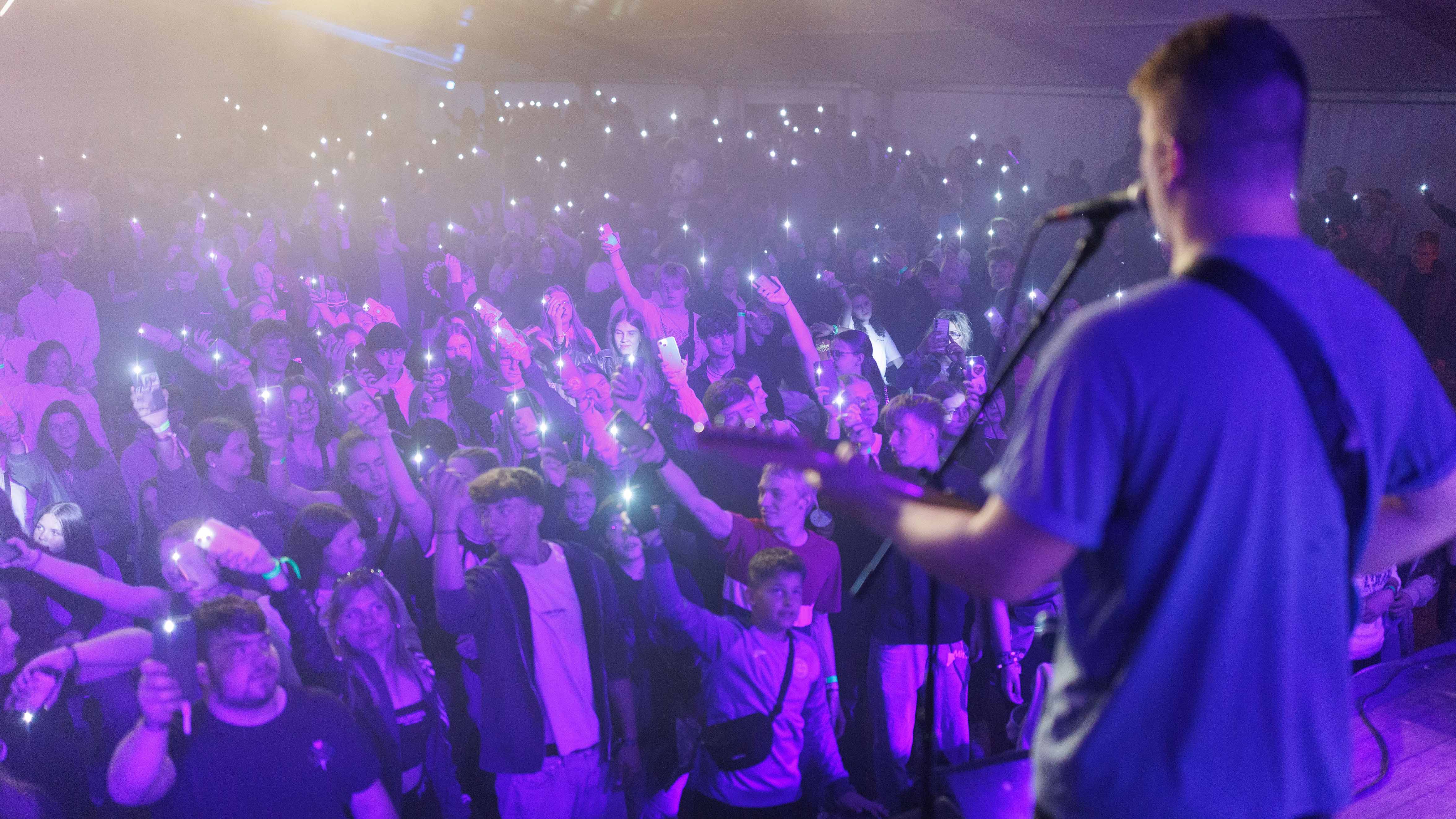 Musiker auf der Bühne singt ins Mikrofon, während ein dichtes Publikum davor steht und viele junge Menschen ihre Smartphones mit eingeschaltetem Licht hochhalten.