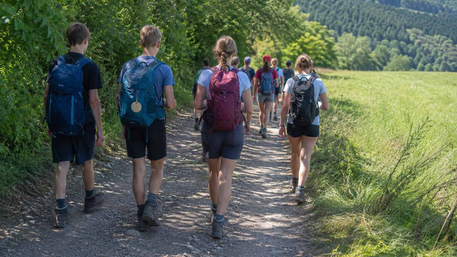 Jugendliche_Pilgerer_Symbolbild_C_KI_ChatGPT_Image_16zu9.jpg Eine Gruppe Jugendlicher mit Rucksäcken wandert auf einem unbefestigten Weg zwischen Bäumen und einer Wiese.