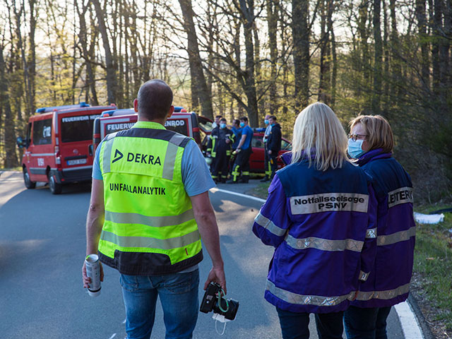 Notfallseelsorger stehen mit lila Jacken vor einem Unfall und einem Feuerwehrauto