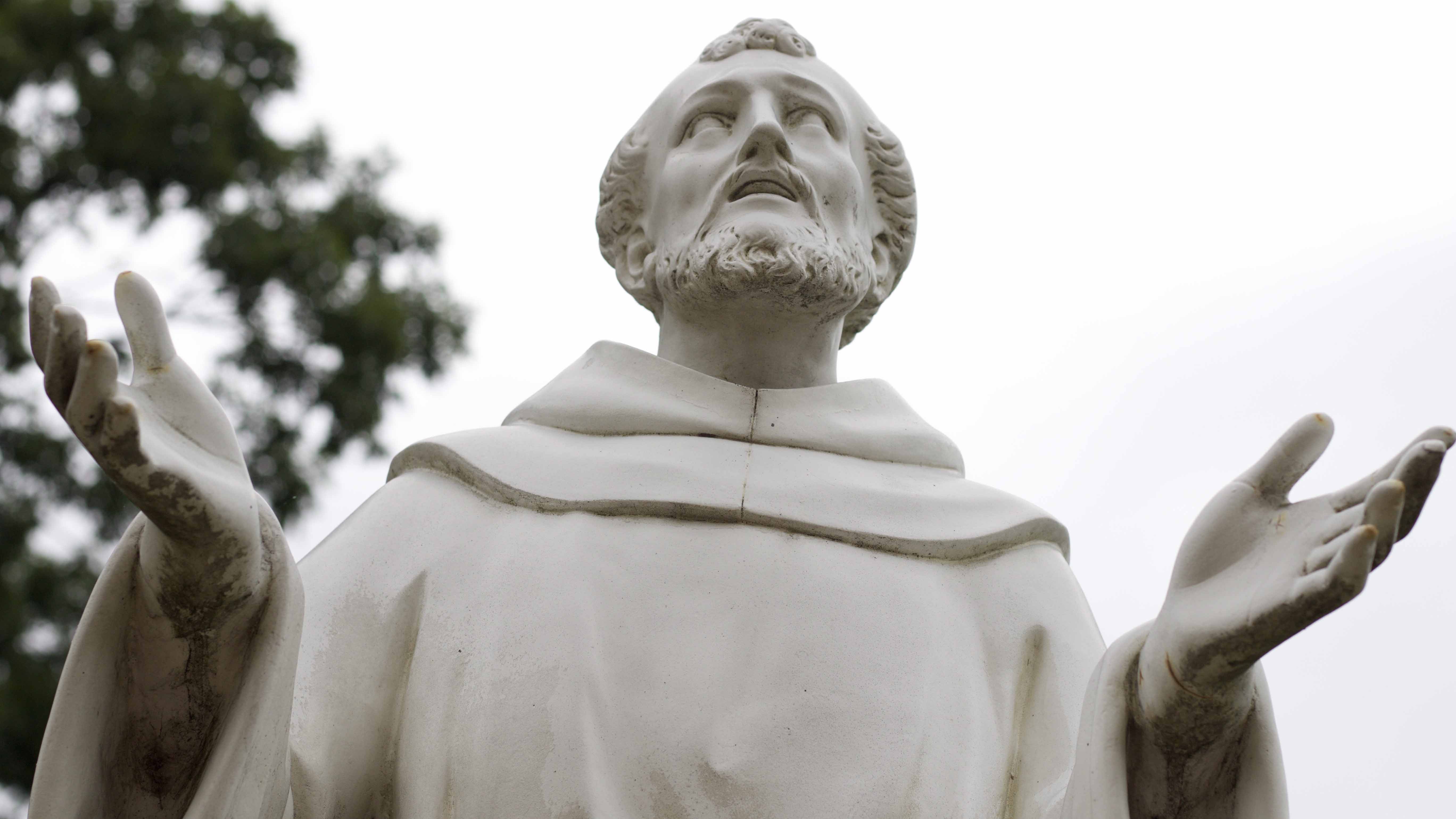 Franz_von_Assisi_C_jdcovell_Pixabay_16zu9.jpg Weiße Statue des heiligen Franz von Assisi in Nahaufnahme, von unten fotografiert. Der bärtige Ordensgründer trägt eine schlichte Mönchskutte, blickt zum Himmel und hat beide Arme mit geöffneten Händen ausgebreitet. Im Hintergrund sind ein bewölkter Himmel und unscharfe Bäume zu sehen.