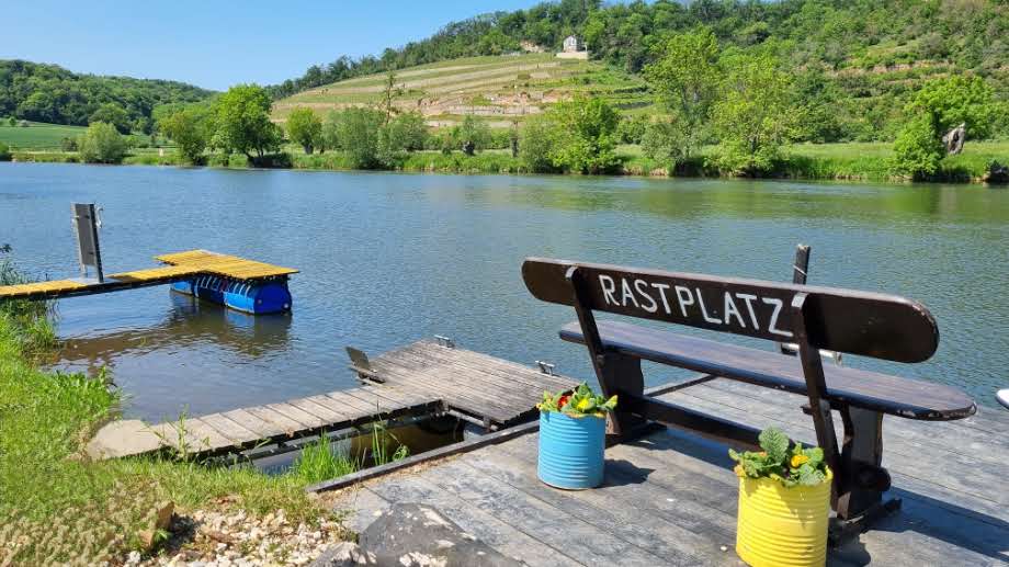 Eine Ruhebank am Flussufer mit der Aufschrift "Rastplatz". vor der Ruhebank stehen ein gelber und ein blauer Pflanzkübel mit Blumen. Auf der gegenüberliegenden Seite des Flusses sind Bäume und grüne Sträucher zu erkennen. Im Hintergrund ein wolkenloser, blauer Himmel.
