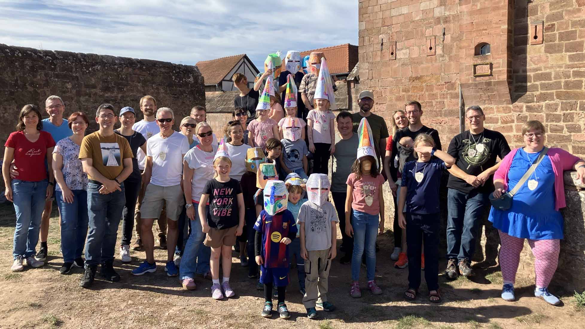 **Alternativtext:**   Gruppenfoto einer Familienfreizeit vor dem Hauptportal der Burg Breuberg. Erwachsene und Kinder stehen gemeinsam vor der historischen Burgmauer. Mehrere Kinder tragen selbst gebastelte, bunt bemalte Ritterhelme und hohe Hüte aus Papier.