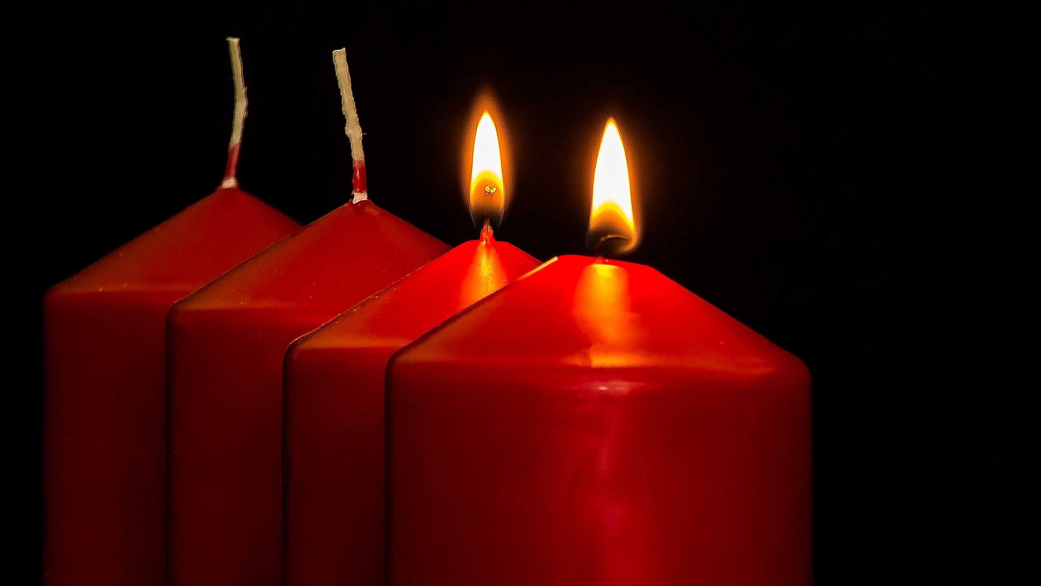 Vier rote Adventskerzen vor schwarzem Hintergrund. Zwei Kerzen brennen, zwei sind noch nicht angezündet. Die Flammen tauchen die Kerzen in warmes Licht.