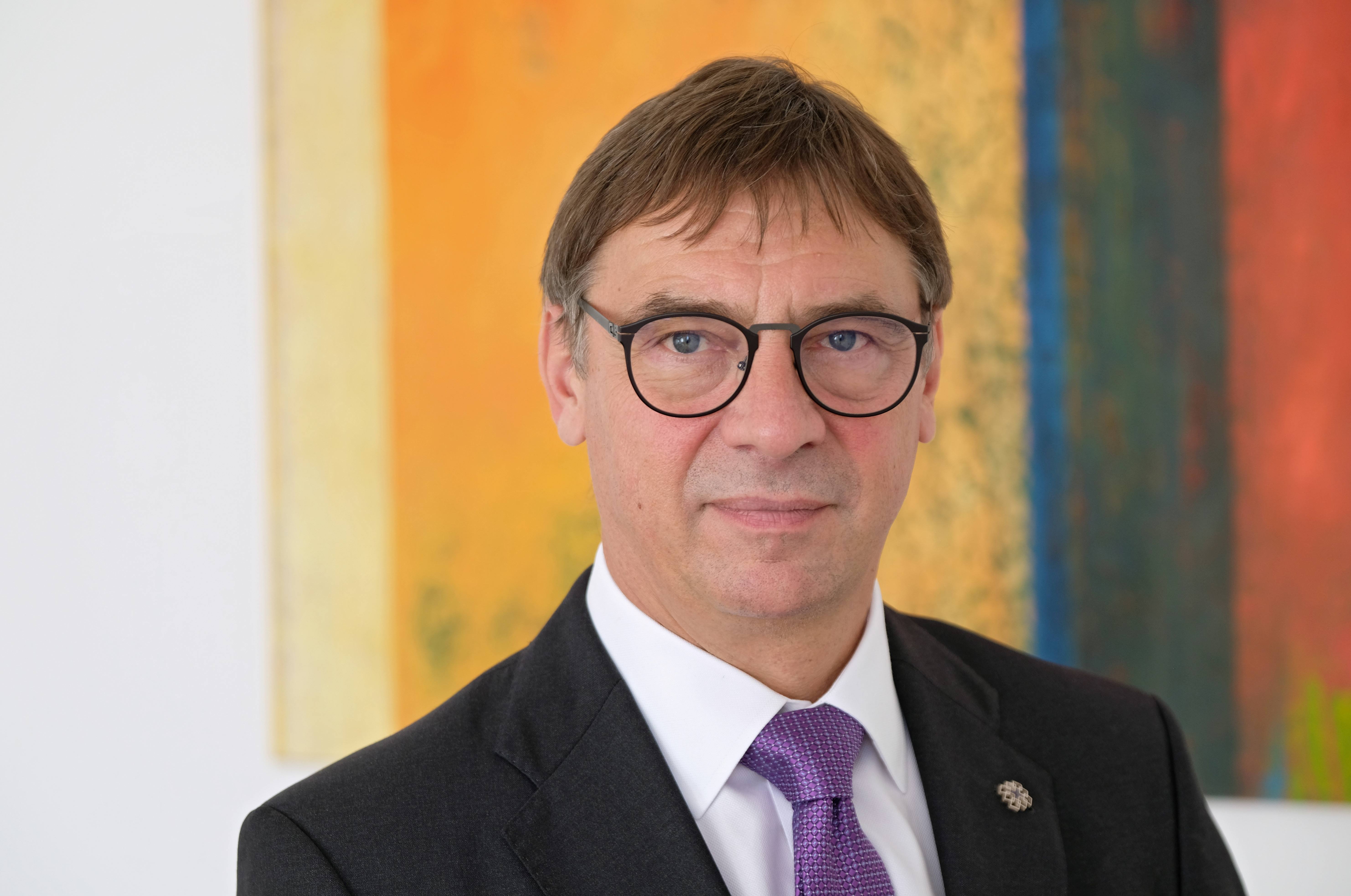 Volker Jung, im dunklen Anzug mit violetter Krawatte und mit Brille vor einem bunten, textilen Wandbehang.