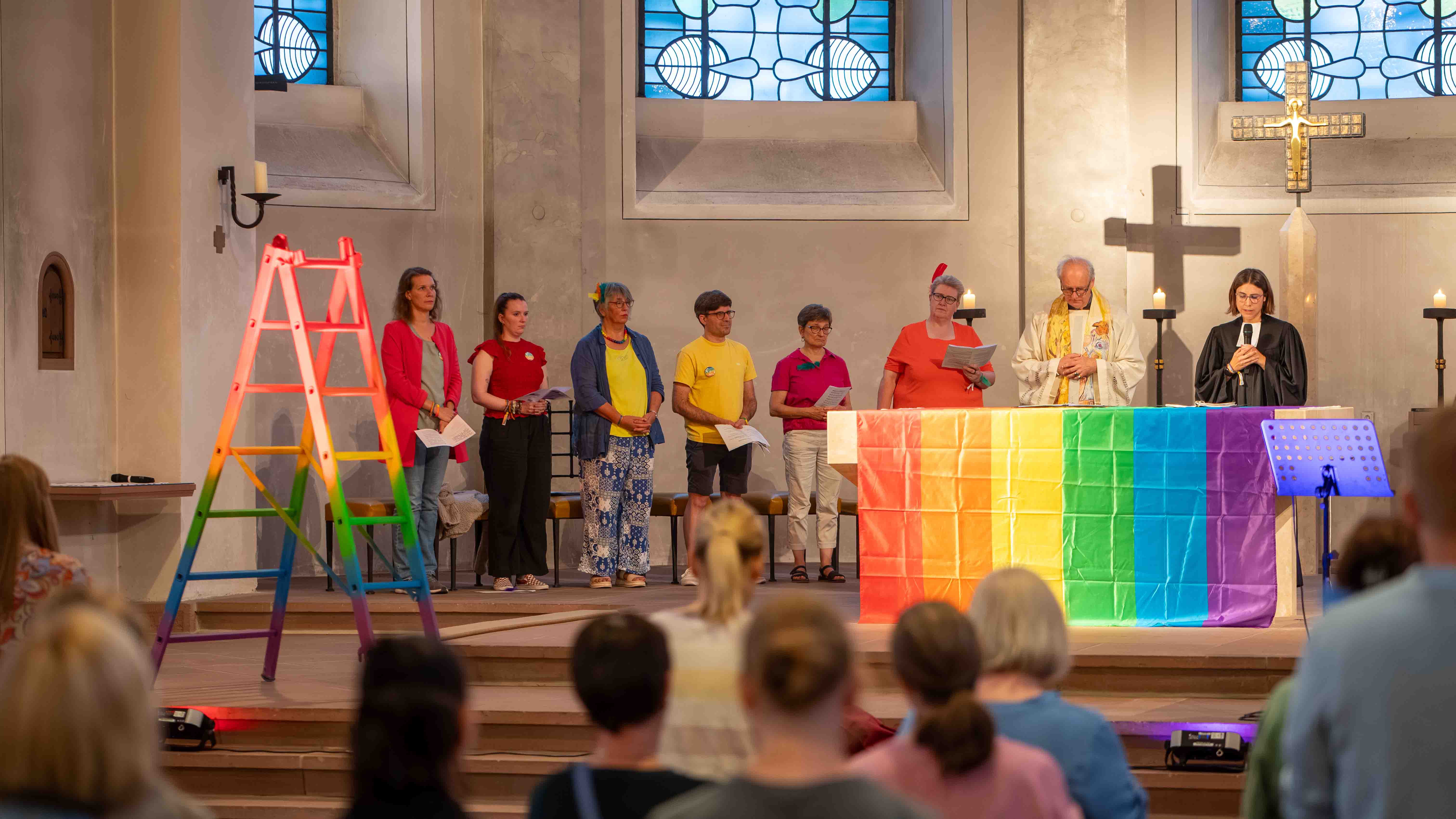Menschen stehen bei einem Gottesdienst vor einem Altar mit Regenbogenfahne; daneben steht eine regenbogenfarbene Leiter.