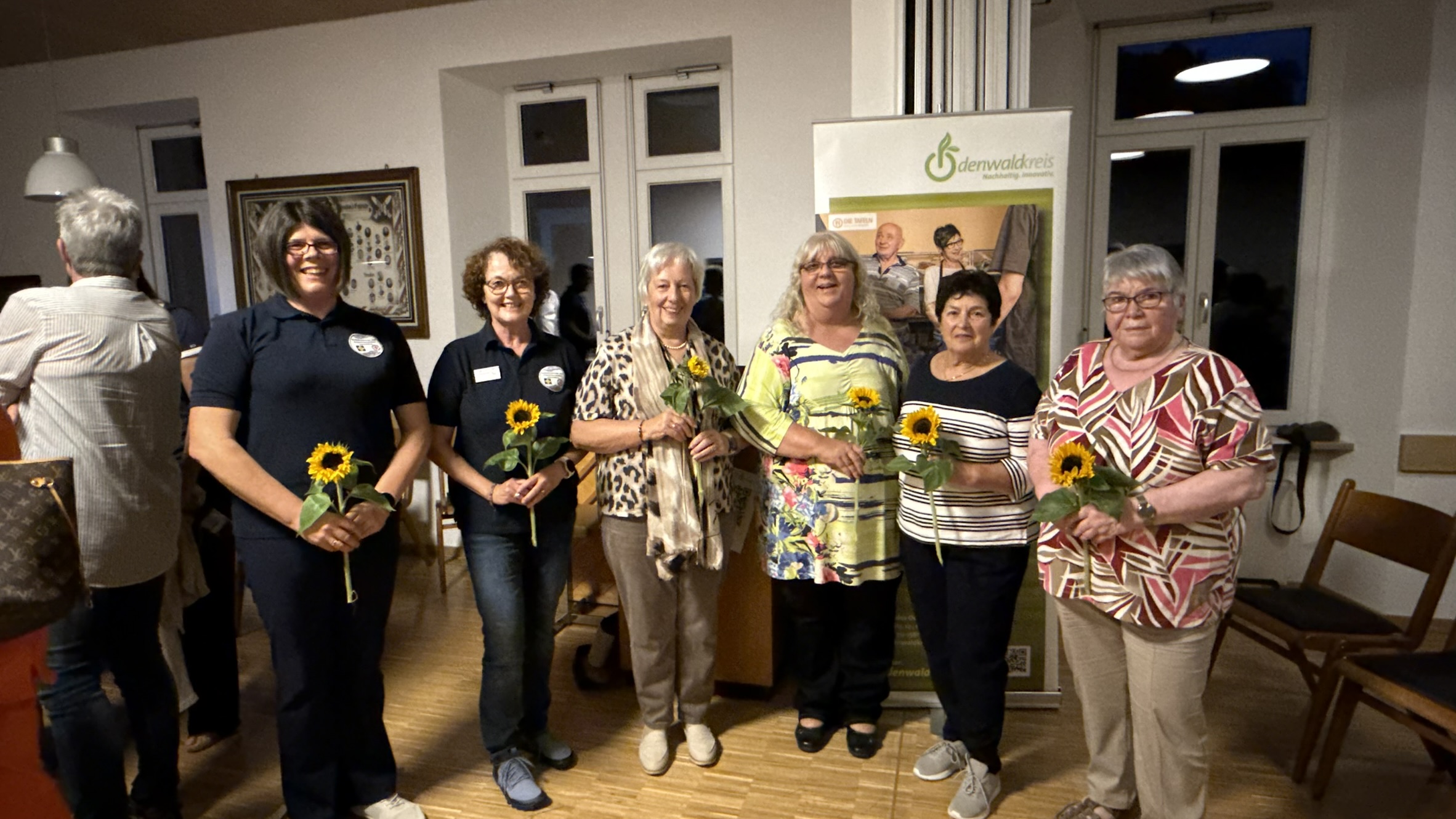 Fünf Frauen, die Sonnenblumen in ihren Händen halten, stehen in einem festlich anmutenden Rahmen in einer Reihe