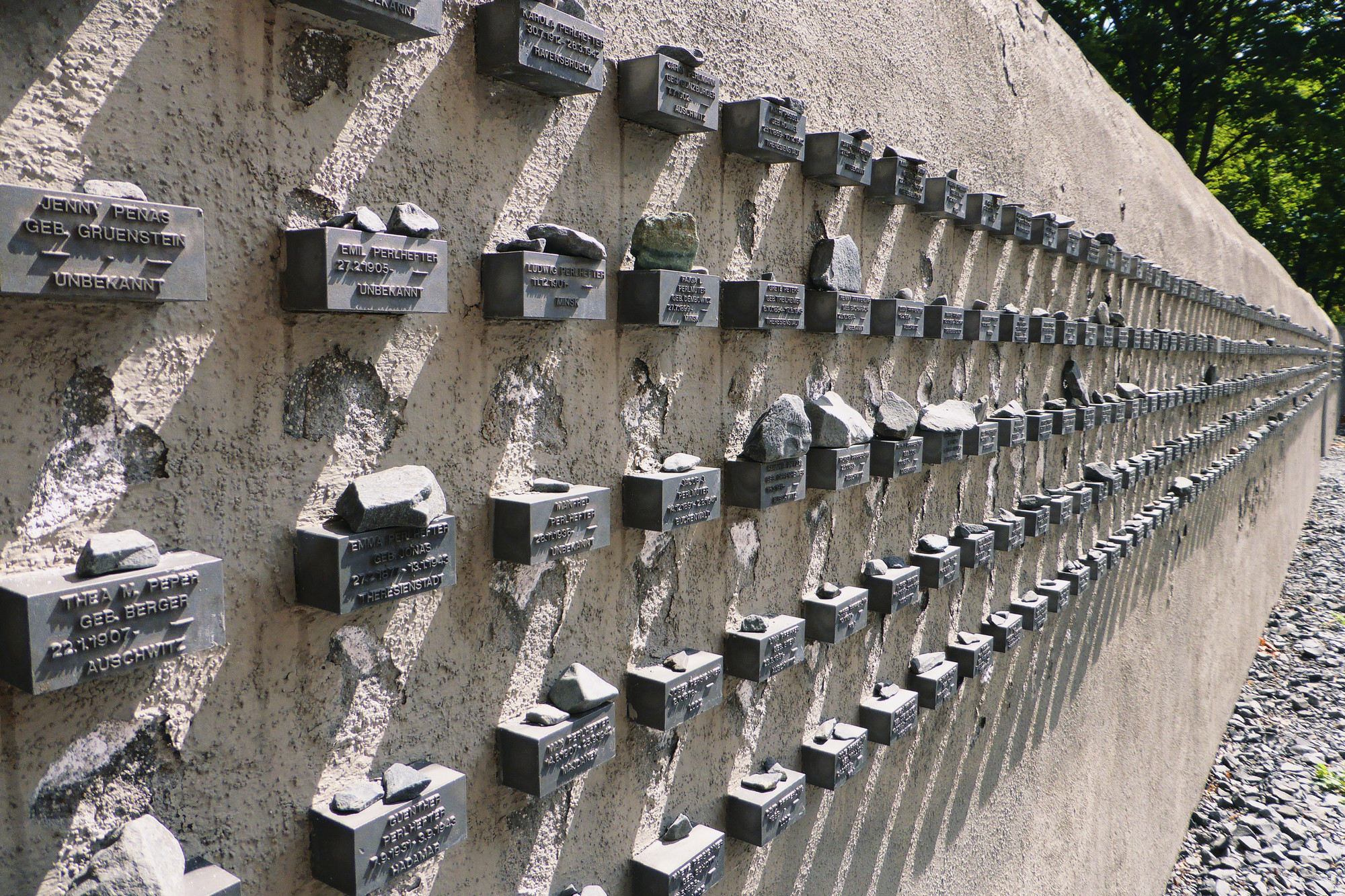 Zu sehen sind Gedenktafeln an der Mauer des jüdischen Friedhofs Battonstraße in Frankfurt/M für die aus der Stadt deportieren jüdischen Menschen. Seitlich an der Mauer befinden sich kleine Boxen, in welche Namen sowie Geburts- und Todesdaten eingraviert sind. Auf diesen Boxen liegen kleine Gedenksteine.