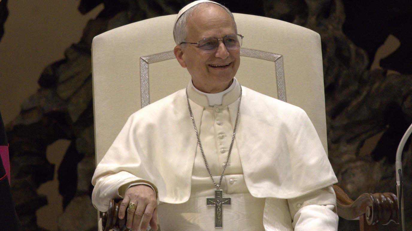 Ein lächelnder Papst in weißem Gewand sitzt auf einem hellen Sessel und trägt ein Brustkreuz an einer langen Kette.