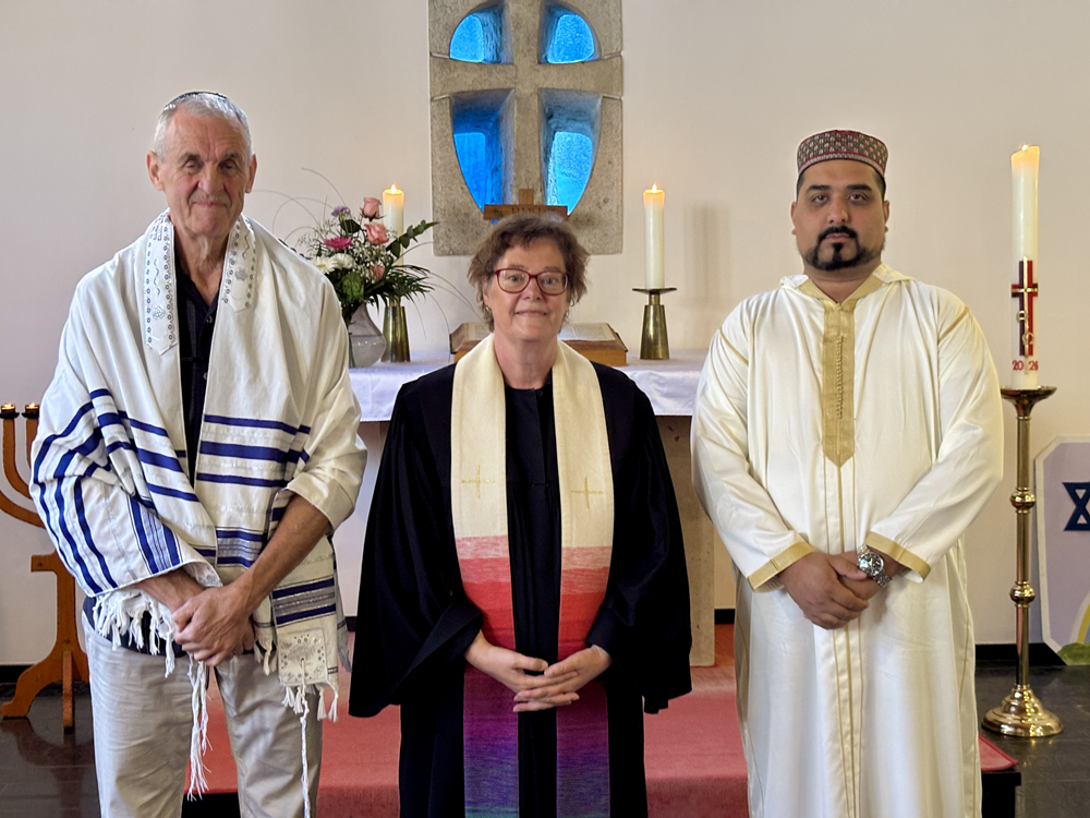 ein jüdischer Geistlicher, eine evangelische Pfarrerin und ein islamischer Imam in ihren gottesdienstlichen Gewändern stehen vor einem Altar