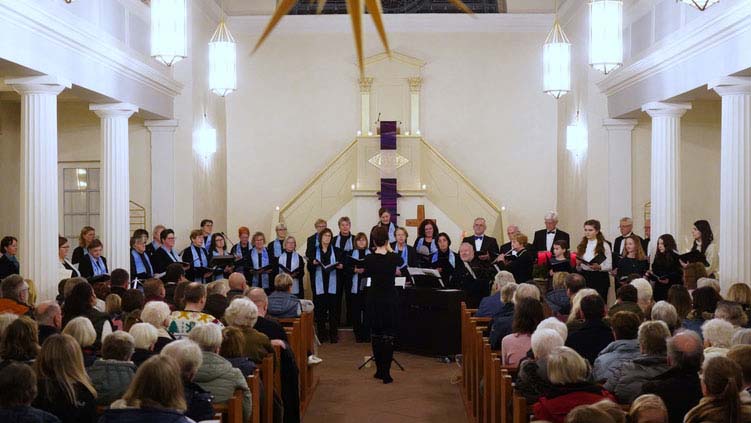 Chor singt unter der Leitung einer Dirigentin vor zahlreichem Publikum in einer hellen Kirche.