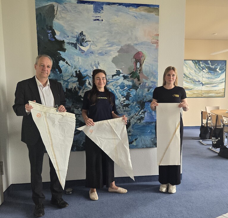 Ein Mann und zwei Frauen stehen nebeneinander und halten jeweils ein Stück weißes Segeltuch fest.Tpck wei