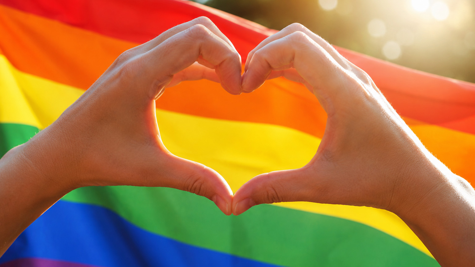 Zwei Hände formen vor einer Regenbogenflagge ein Herz als Symbol für Liebe, Vielfalt, Akzeptanz und Solidarität.