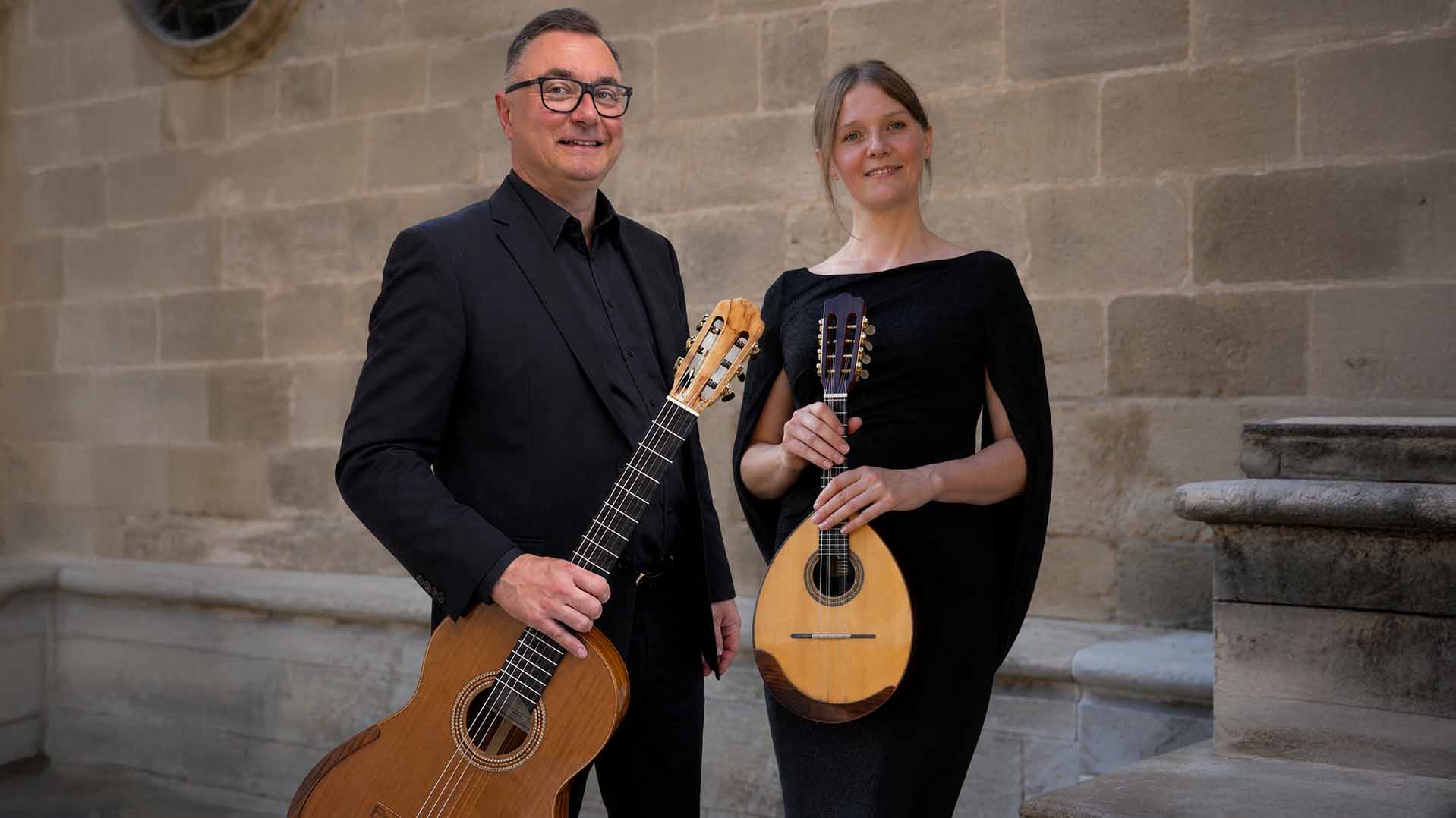 Zwei Konzertmusizierende in schwarzer Kleidung stehen mit Gitarre und Mandoline vor einer Steinmauer und blicken lächelnd in die Kamera.
