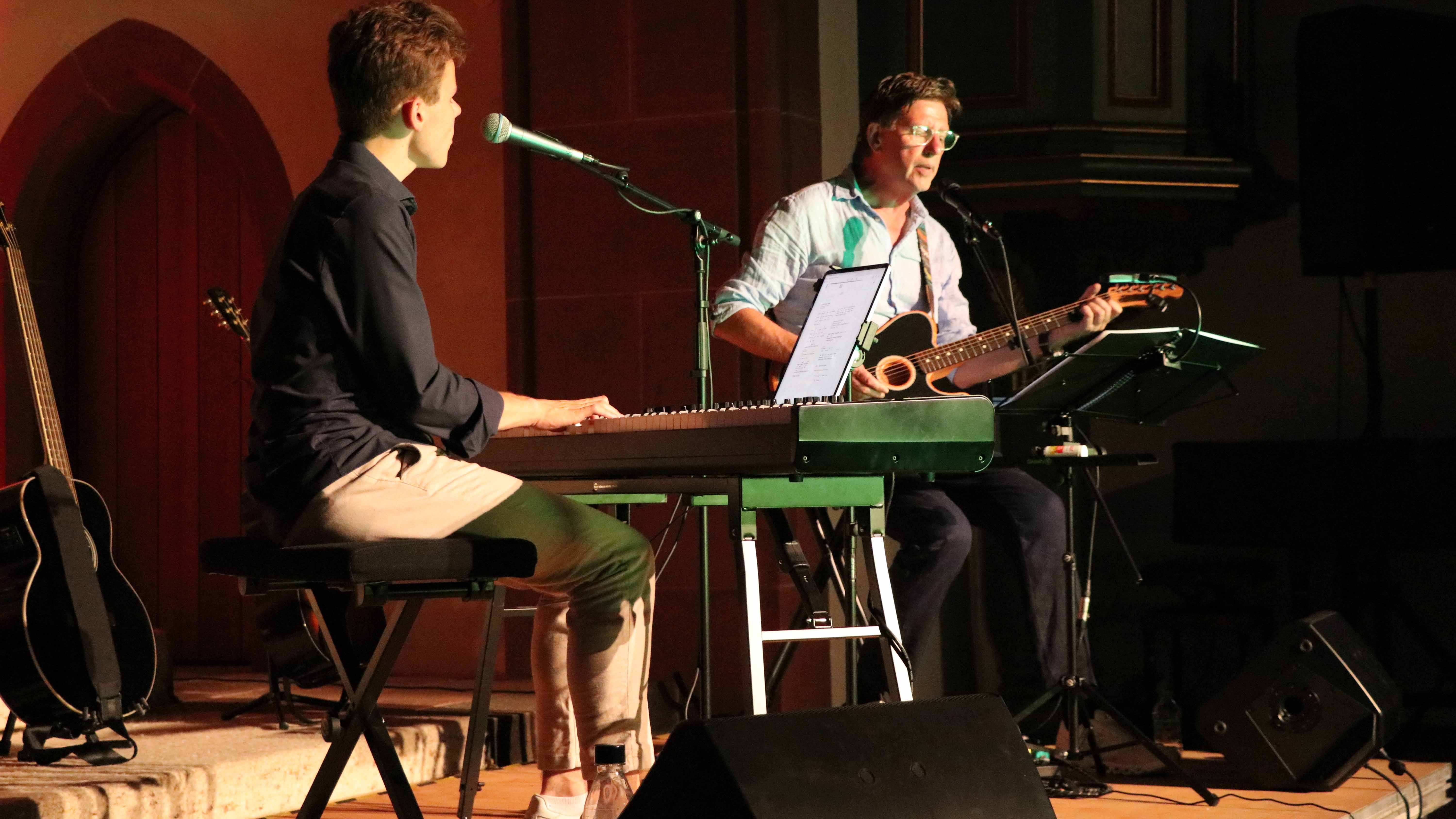 Peter Paletta am E-Piano und Lars Hansen mit Akustikgitarre und Gesang bei einem gemeinsamen Konzert in einer Kirche. Beide Musiker sitzen vor Mikrofonen auf einer stimmungsvoll beleuchteten Bühne.