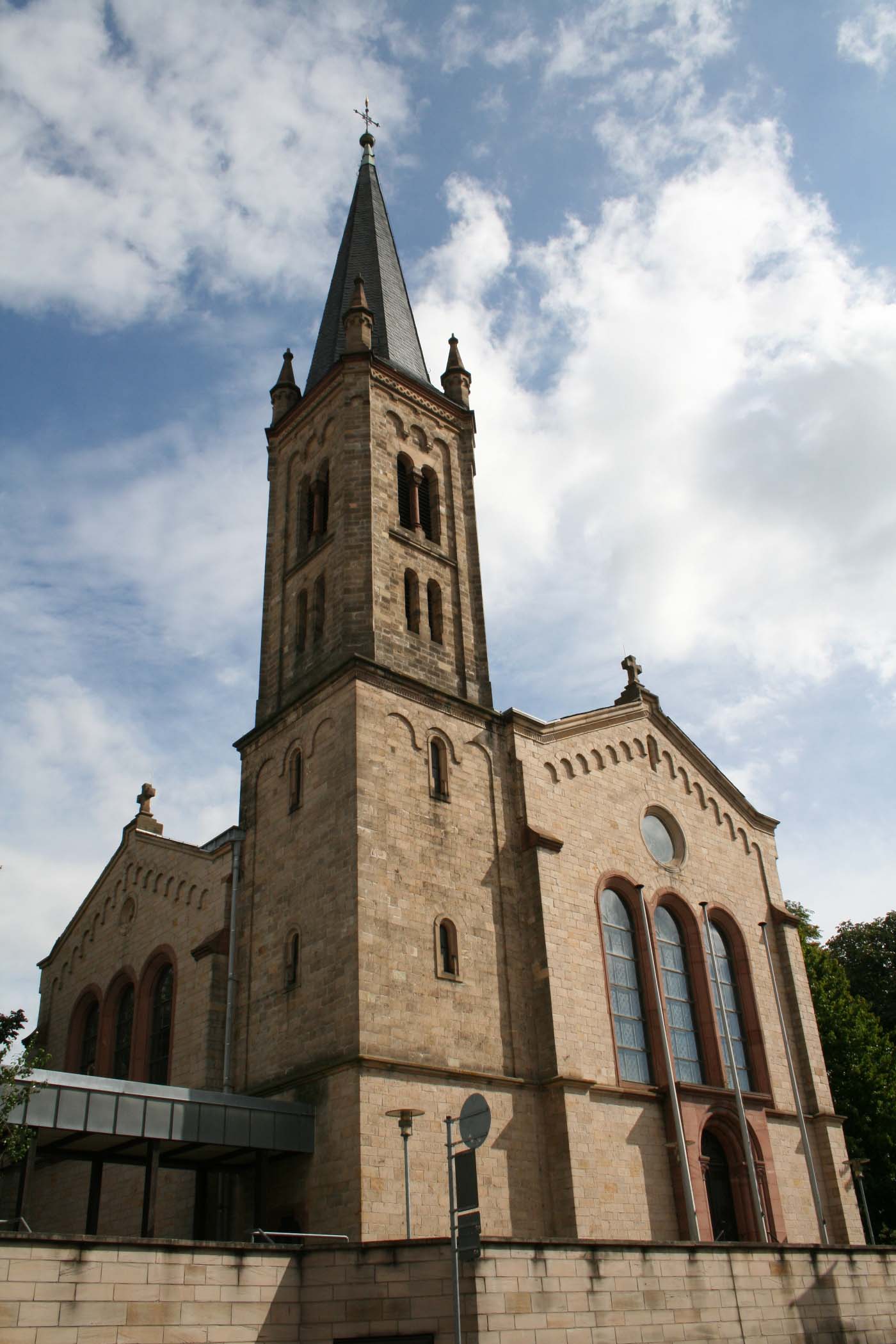 Historische Kirche aus hellem Stein mit hohem Turm und spitzem Dach, gesehen von unten vor bewölktem Himmel.