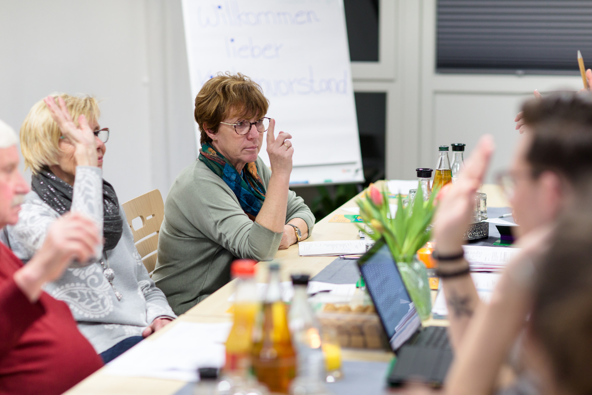 Männer und Frauen sitzen zusammen an einem Tisch und heben zur Abstimmung ihre Hand. Im Fokus des Bildes ist eine Frau zu sehen, welche konzentriert nach vorne schaut. Auf dem Tisch befindet sich eine Vielzahl an Schreibutensilien sowie Kekse und Getränke.