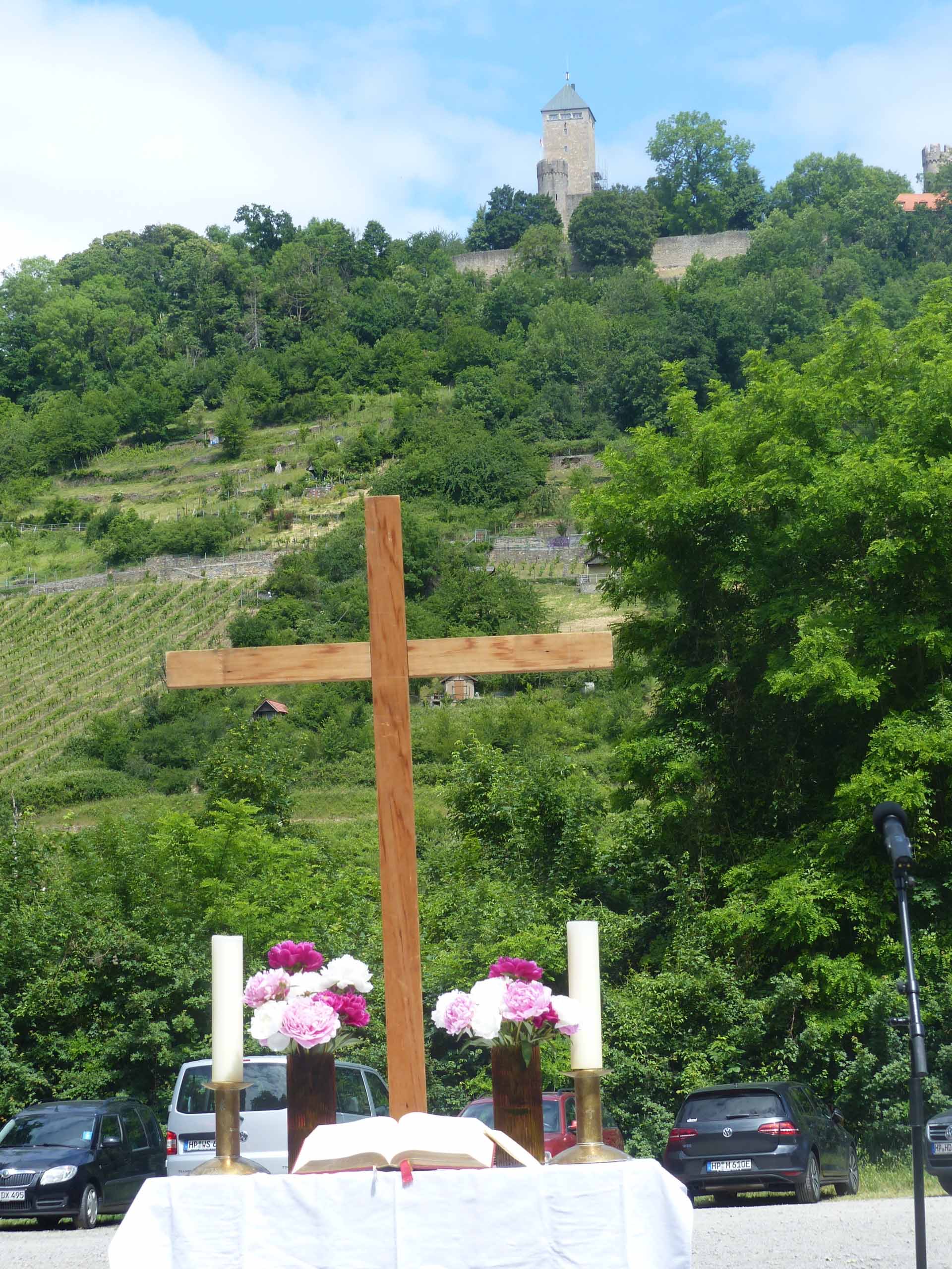 Altar mit Holzkreuz, Kerzen und Blumen vor bewaldetem Hang mit Weinbergen und Burganlage im Hintergrund bei einem Freiluftgottesdienst.