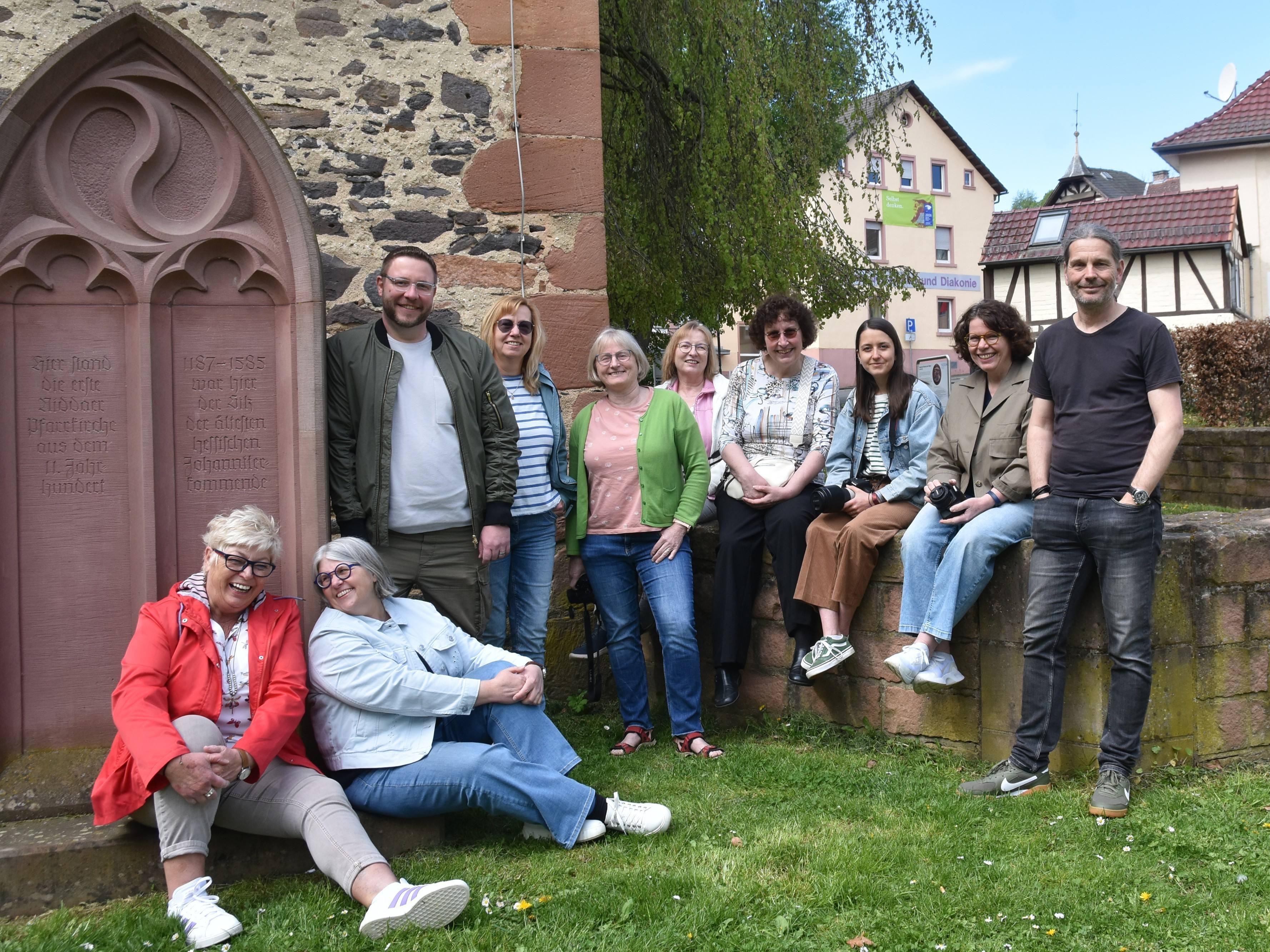 eine Gruppe von zehn Personen, zwei Männer, acht Frauen, hat sich am Johanniter-Turm gruppiert, einige sitzen auf einer Mauer, einige stehen links und rechts von den Sitzenden, zwei Frauen haben sich auf einer Sandsteinplatte vor einem großen alten Grabstein niedergelassen, alle sind bester Laune