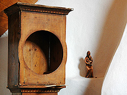Eine leere hölzerne Standuhr, daneben an der Wand eine Marienstatuette mit Kind