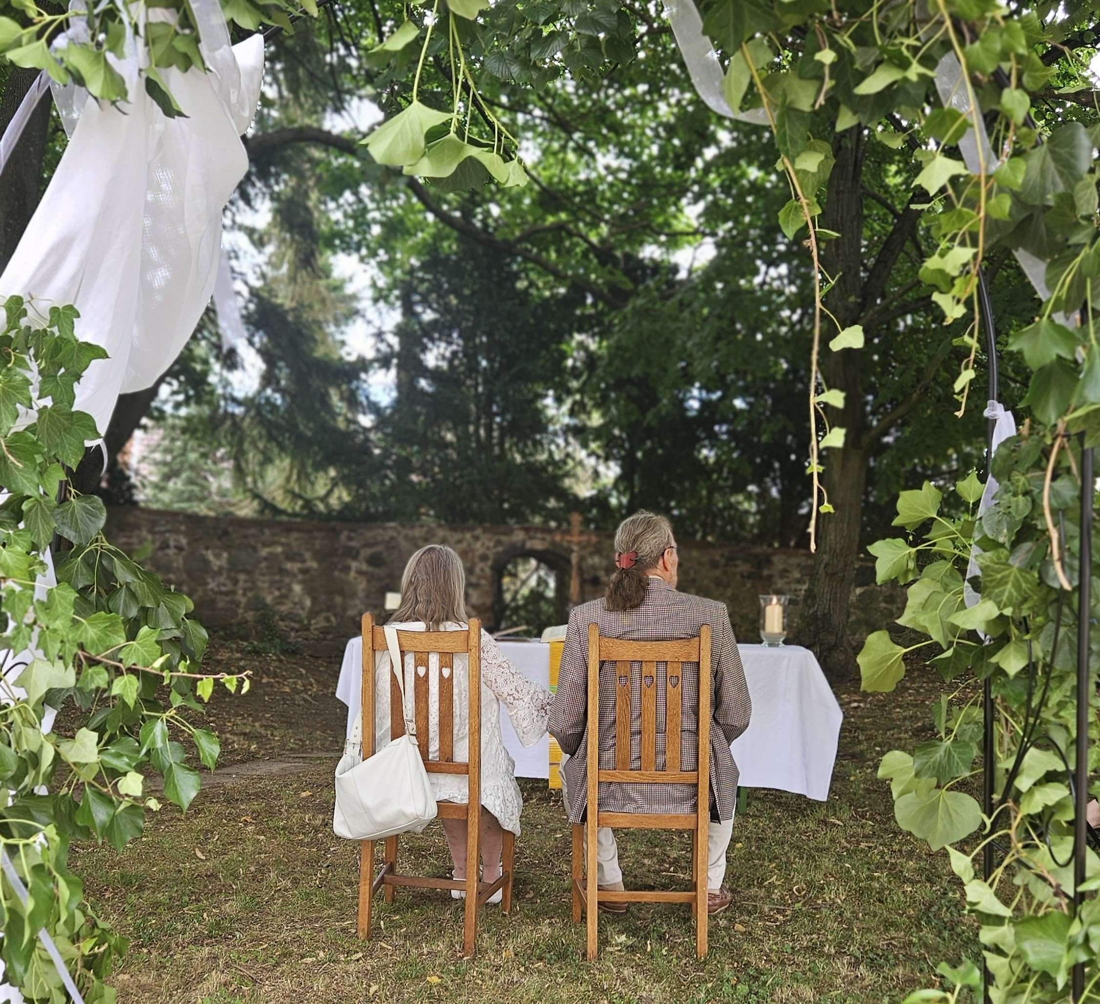 Ein Paar, Ansicht von hinten, sitzt auf Stühlen vor einem improvisierten Altar. Im Vordergrund ein mit Efeuranken und weißen Bändern geschmückter Bogen.