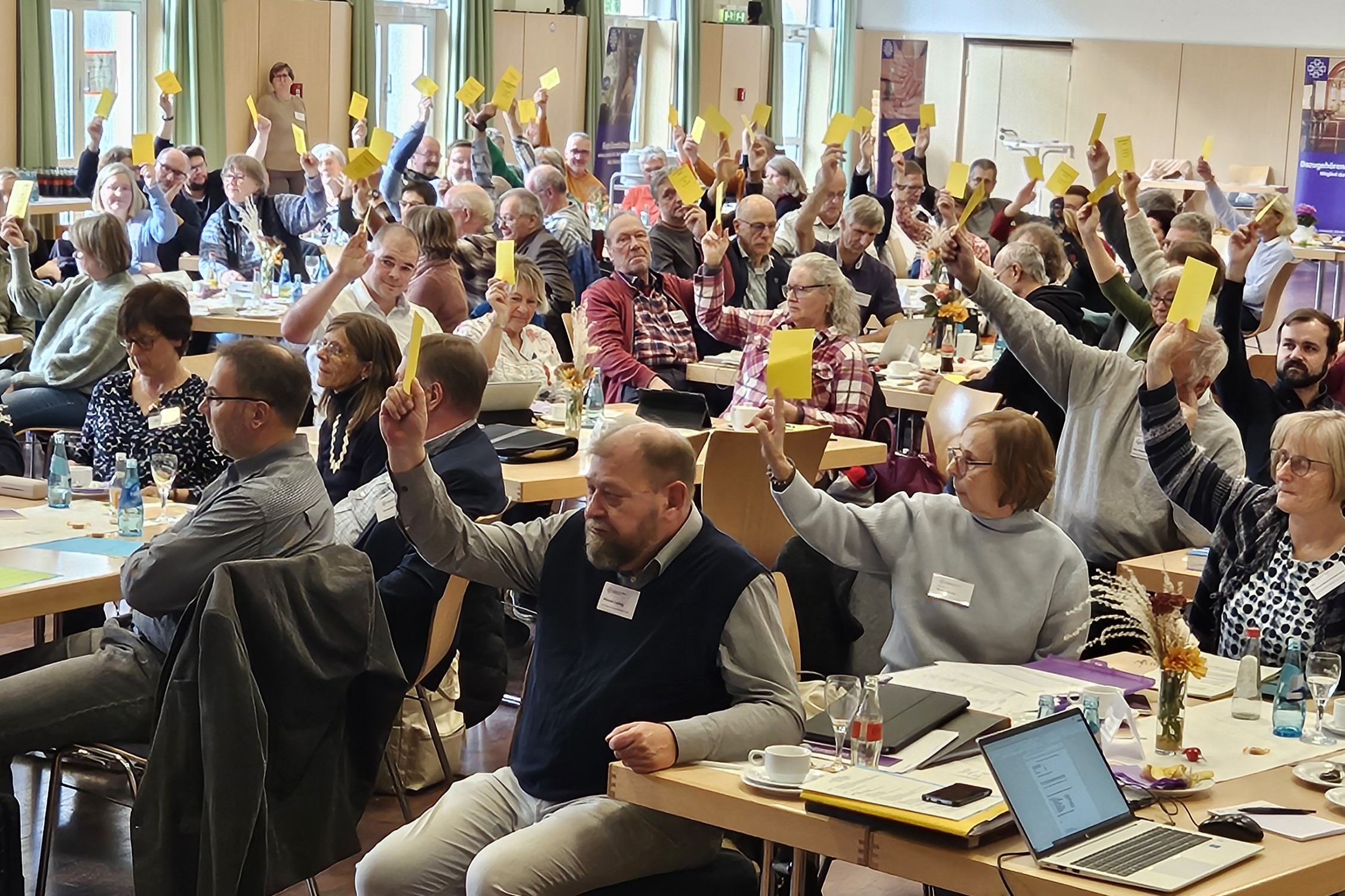 Blick in den Saal des Bürgerhauses Nidda, die Synodalen stimmen ab und halten gelbe Stimmkarten in die Höhe.