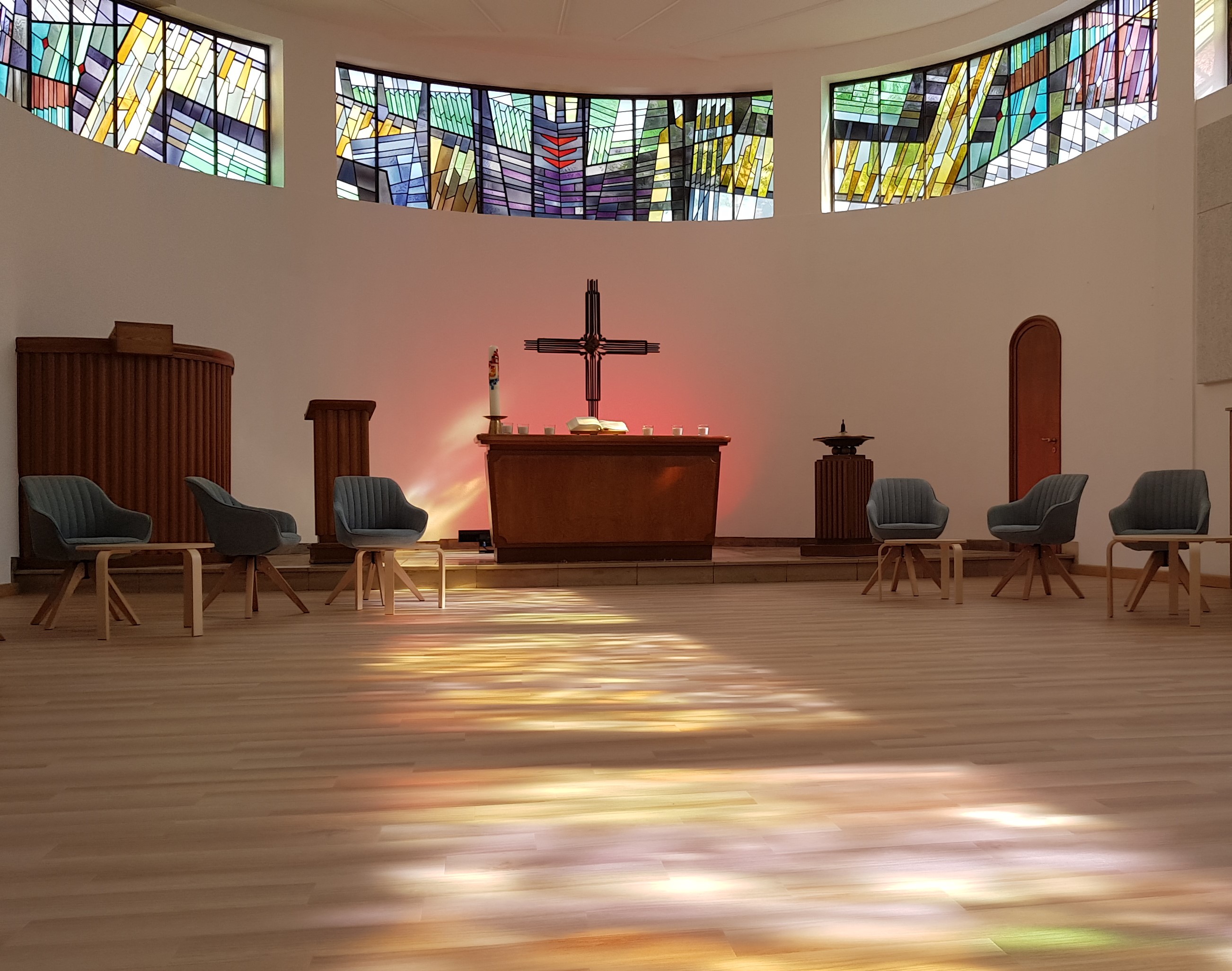 Blick in einen Kirchenraum, durch bunte Fenster fällt sanftes Licht auf den Dielenboden. Statt Bänken gibt es bequeme Stühle.