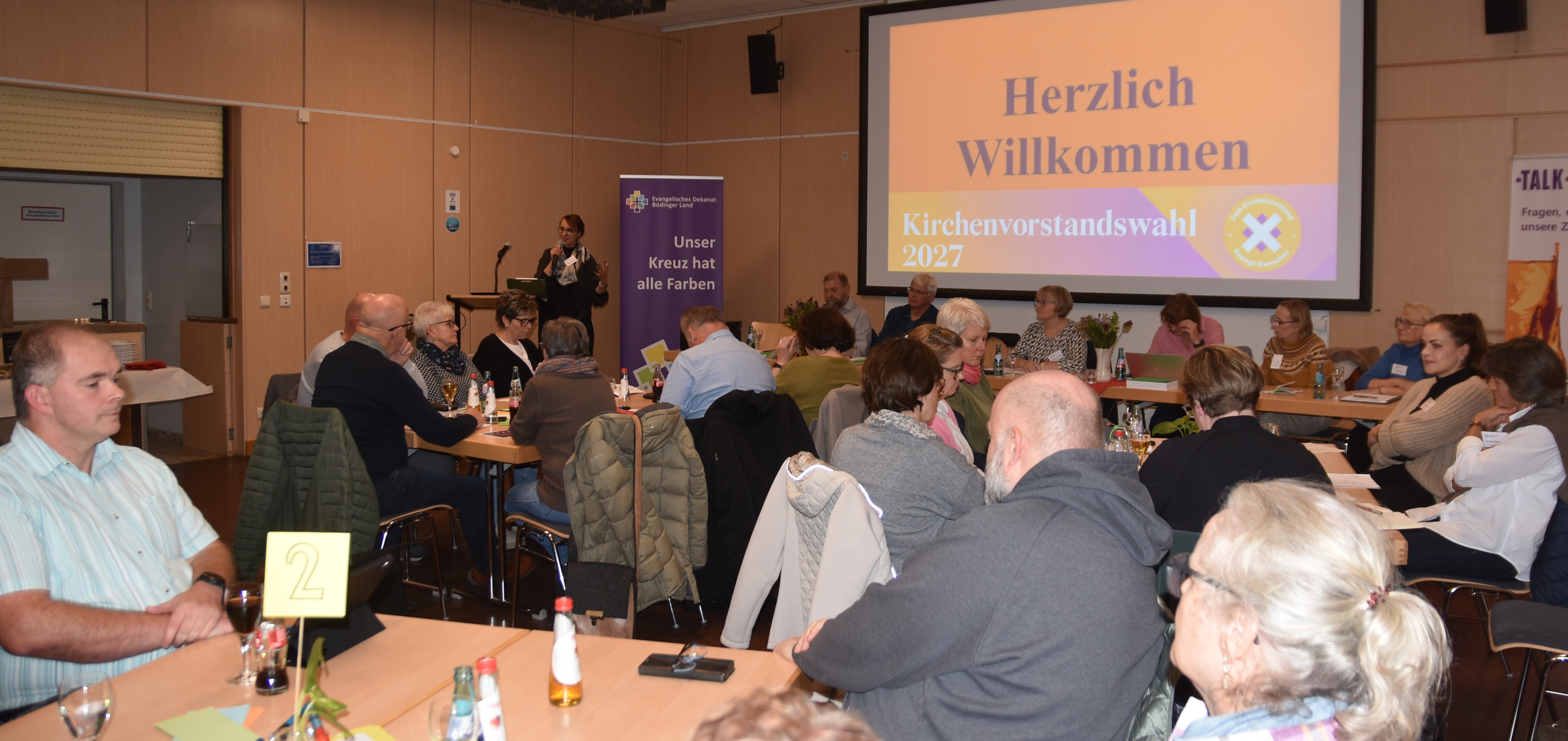 Blick in den vollen kleinen Saal des Bürgerhauses Nidda. Menschen sitzen an Tischgruppen. Vorne steht Dekanin Birgit Hamrich, Mikro in der Hand, und begrüßt. Im Hintergrund auf einem großen Bildschirm steht "Herzlich willkommen". Daneben steht ein Roll-up mit "Unser Kreuz hat alle Farben".