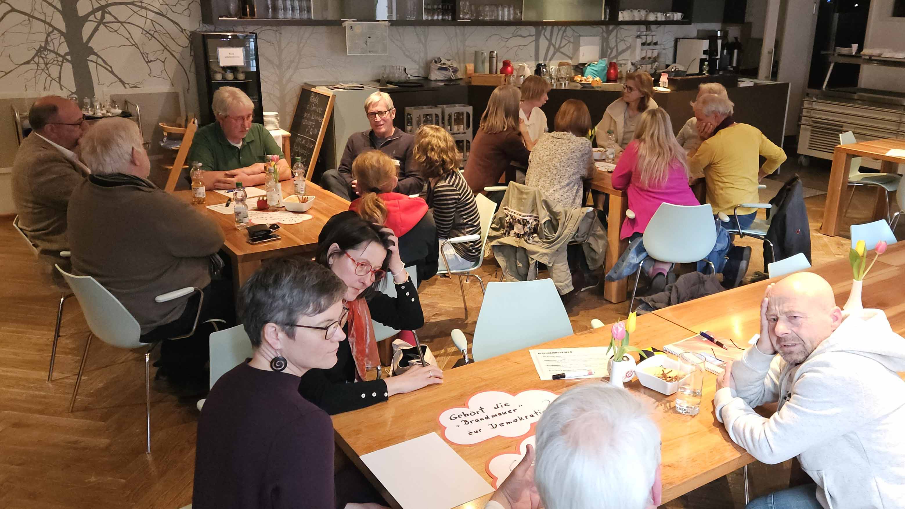 Mehrere Erwachsene sitzen in einem Café an Tischen und diskutieren in kleinen Gruppen; auf den Tischen liegen Notizen und stehen Getränke.