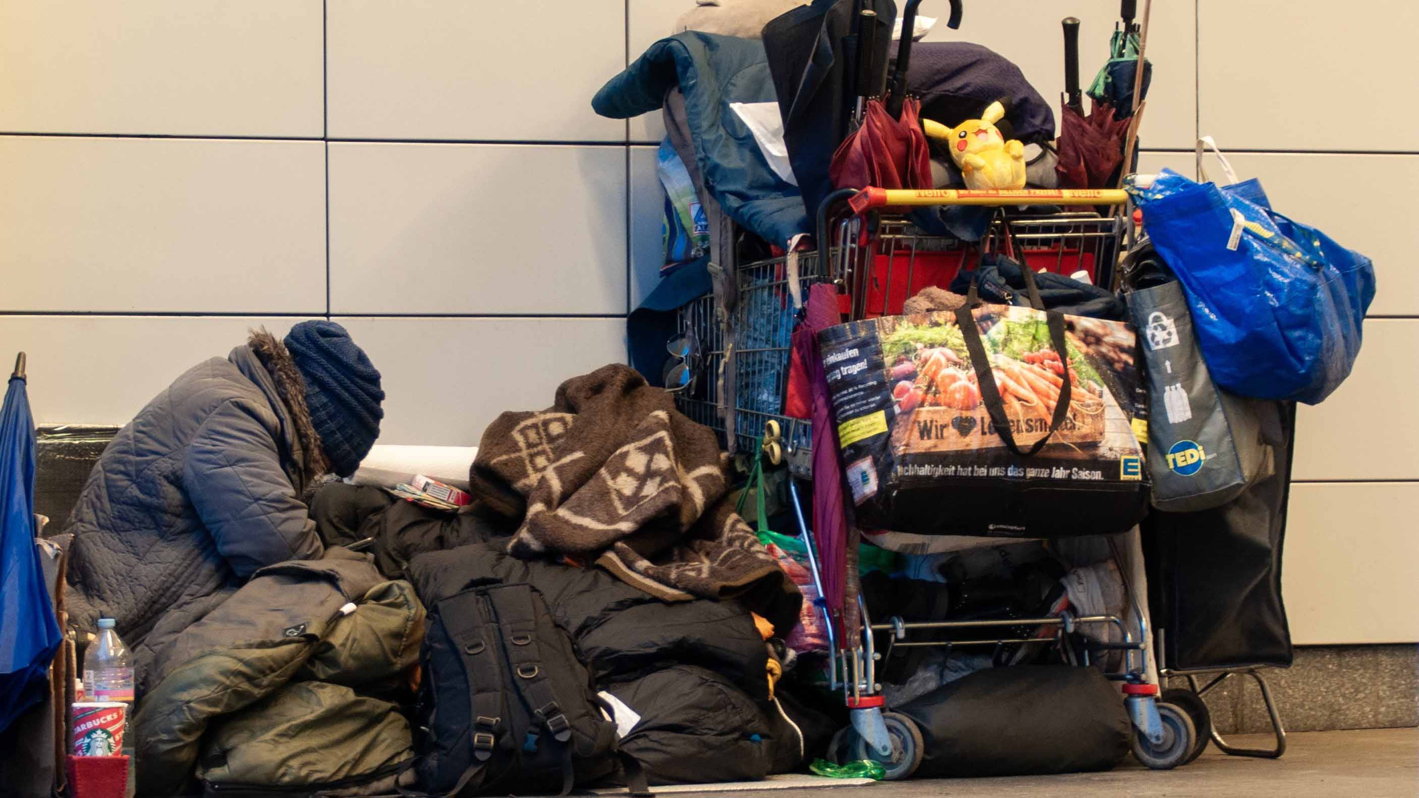 Eine Person sitzt zwischen Gepäck und Decken neben einem voll beladenen Einkaufswagen mit Taschen, Kleidung, Schirmen und persönlichen Gegenständen.
