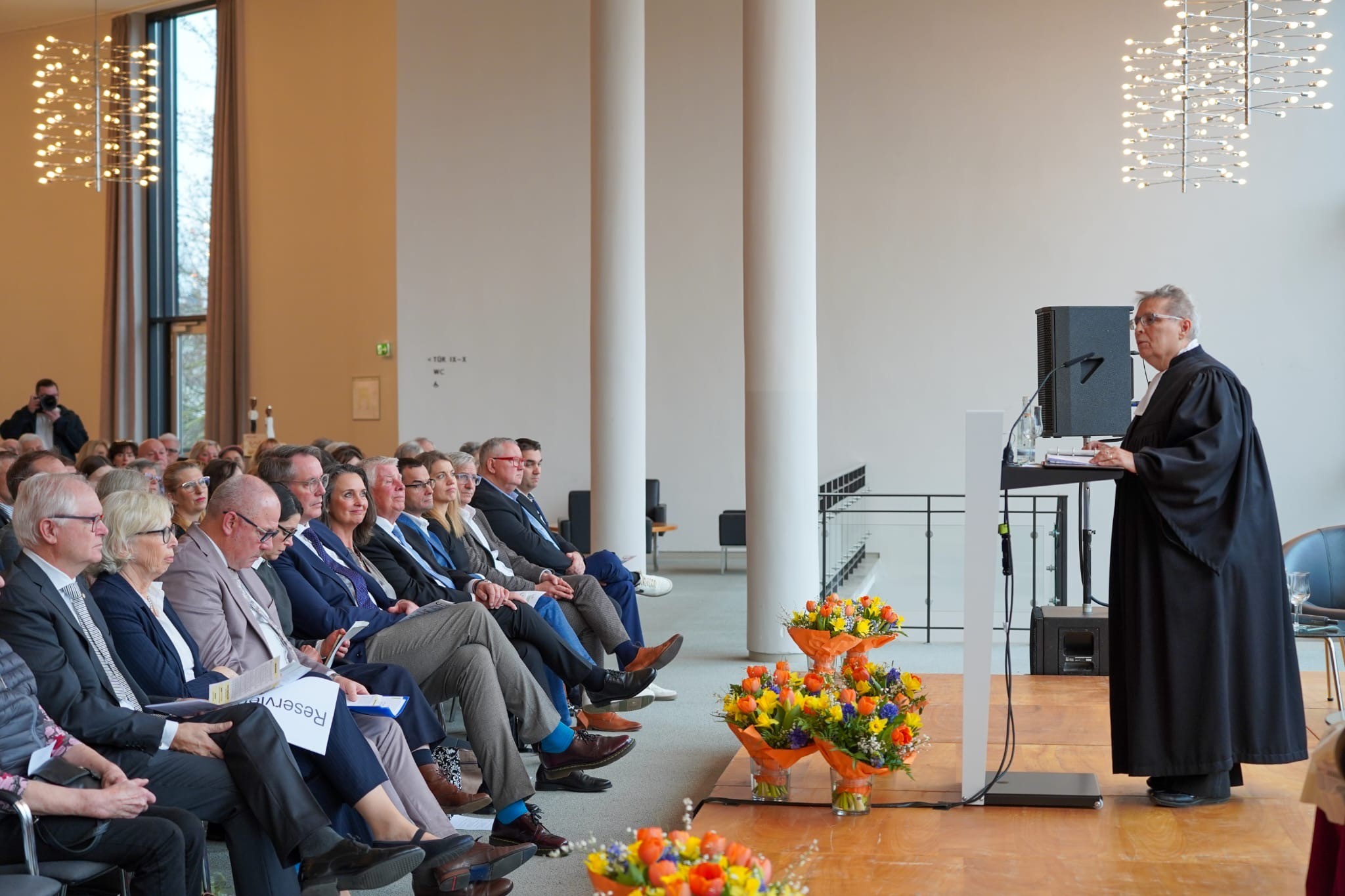 Jubiläumsfeier der Ökumenischen Hospizhilfe Worms Eine Person in Robe hält eine Rede vor einer sitzenden Zuhörerschaft in einem hellen, modernen Saal mit großen Fenstern, Blumenarrangements auf dem Podium und "Reserviert"-Schildern an einigen Stühlen.