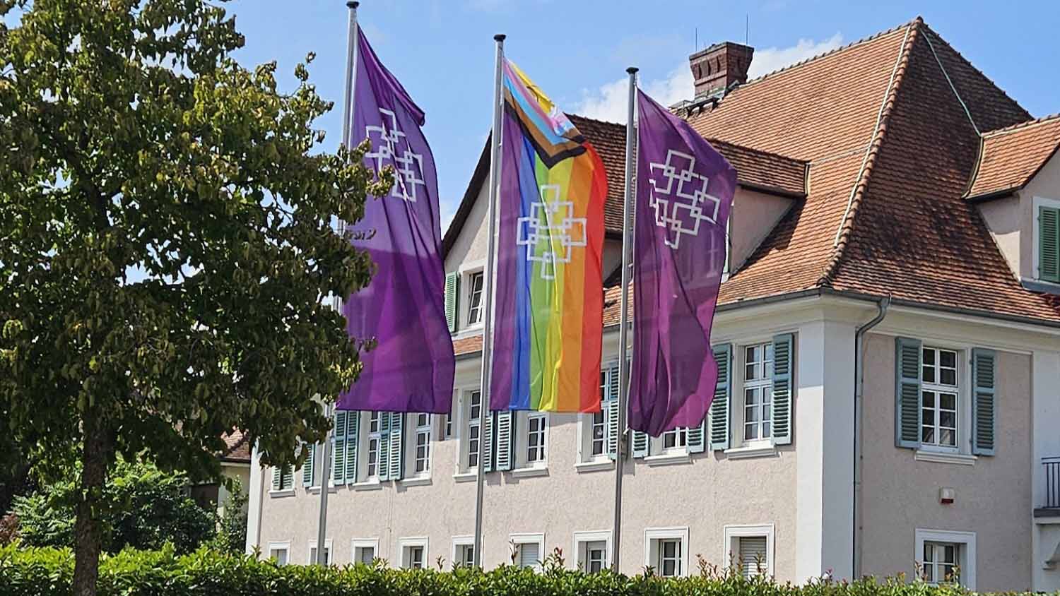 Vor einem hellen Gebäude mit roten Ziegeldächern und grünen Fensterläden, dem Haus der Kirche in Heppenheim, stehen mehrere Fahnenmasten. In der Mitte weht die Progress-Pride-Flagge mit Regenbogenstreifen und zusätzlichen farbigen Winkeln. Daneben hängen zwei violette Fahnen mit einem weißen grafischen Symbol, dem EKHN-Facettenkreuz. Links im Bild steht ein großer Baum, im Vordergrund ist eine Hecke zu sehen.