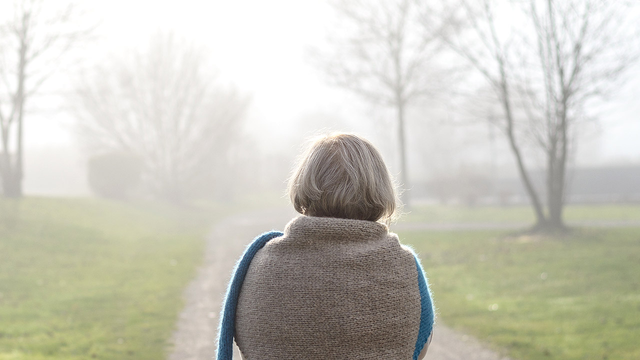 Eine ältere Dame geht spazieren, vor ihr ragen Bäume aus dem Nebel