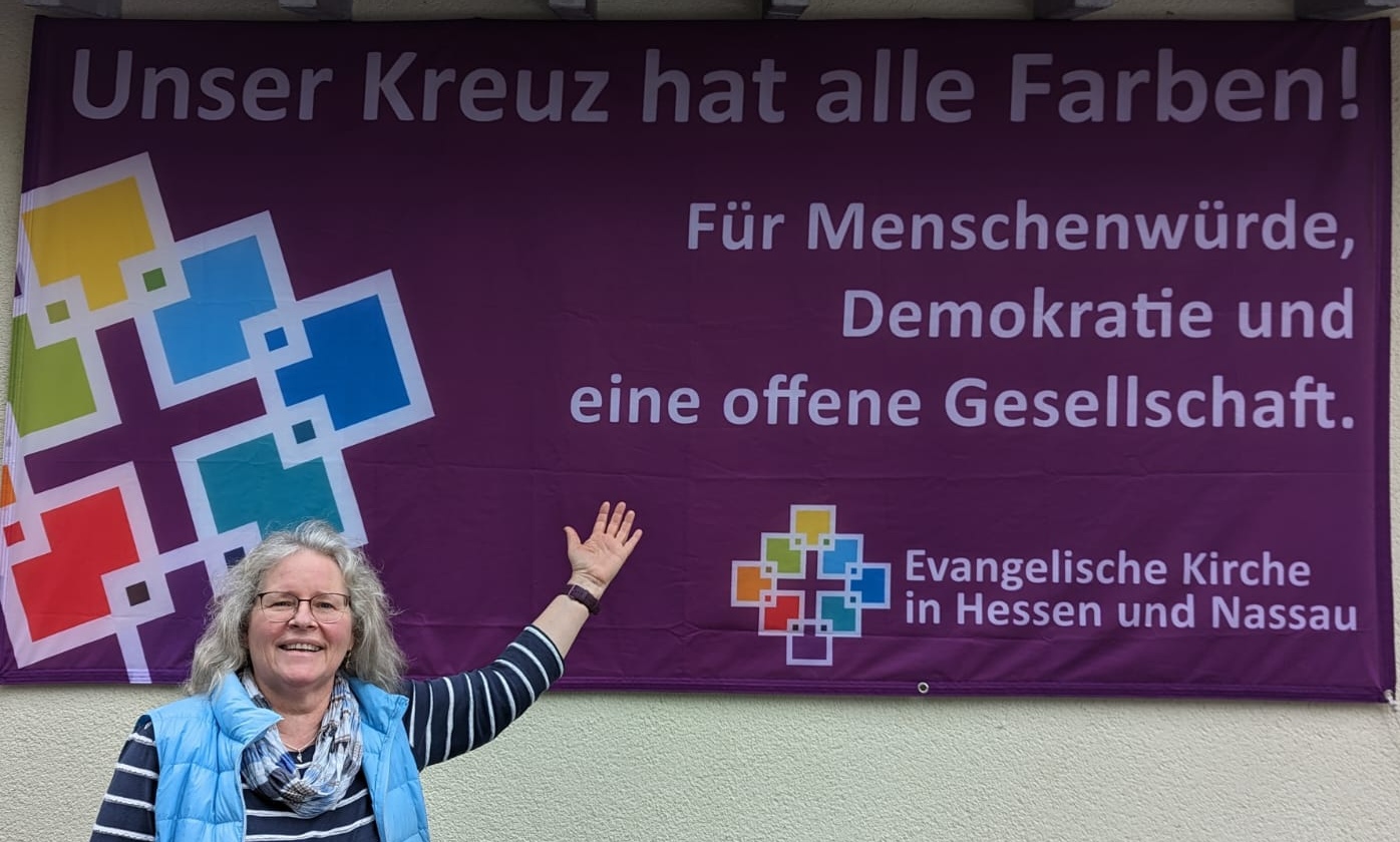 Pfarrerin Hanne Allmansberger zeigt vor dem Niddaer Johannes-Pistorius-Haus auf das lilafarbene Querbanner der Aktion, das dort aufgehängt wurde.
