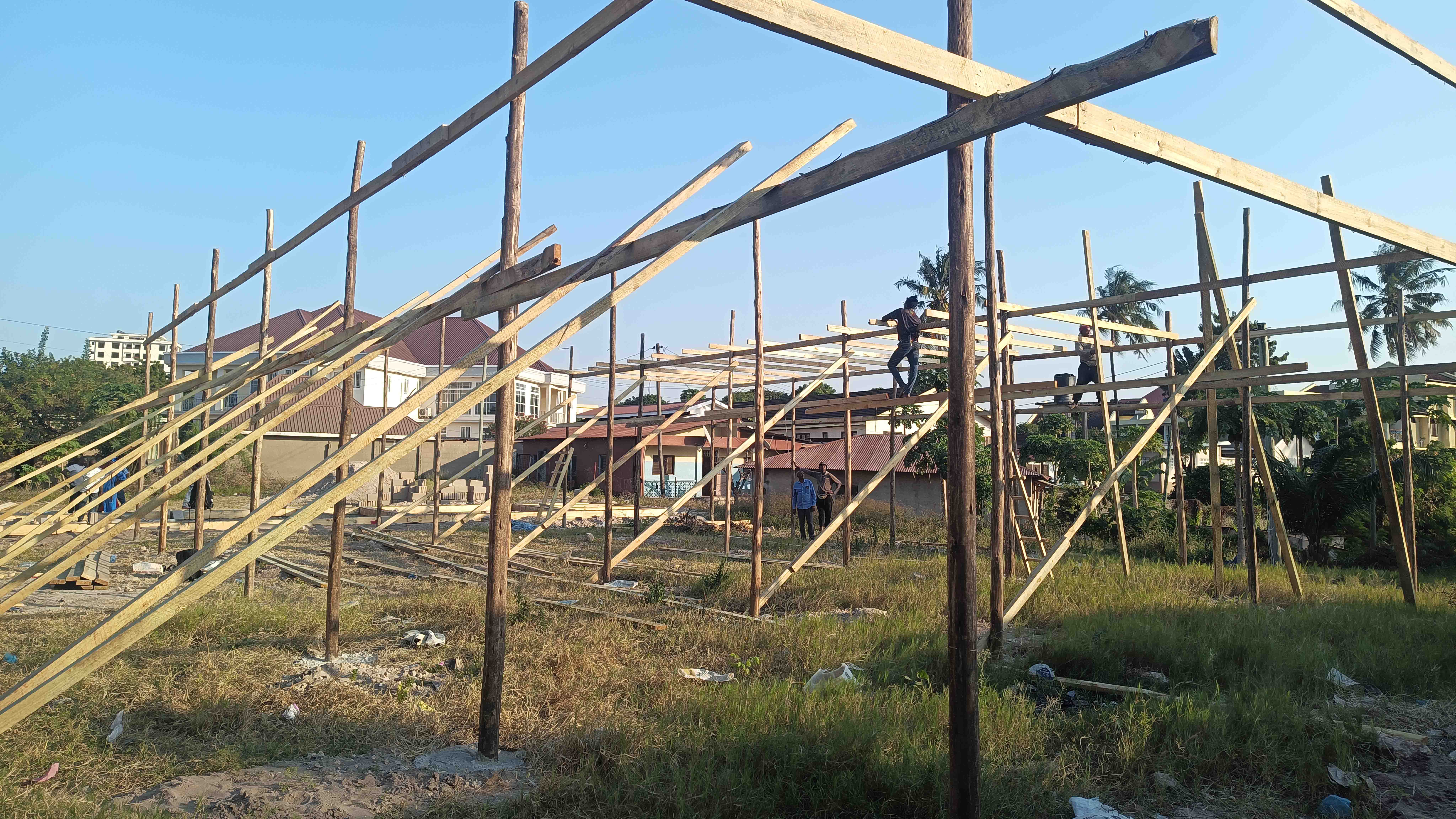 Mehrere Arbeiter errichten auf einem freien Gelände eine provisorische Holzkonstruktion. Senkrechte Pfosten, Querbalken und schräge Verstrebungen bilden bereits das Gerüst der Behelfskirche. Im Hintergrund sind Häuser und Palmen zu sehen.