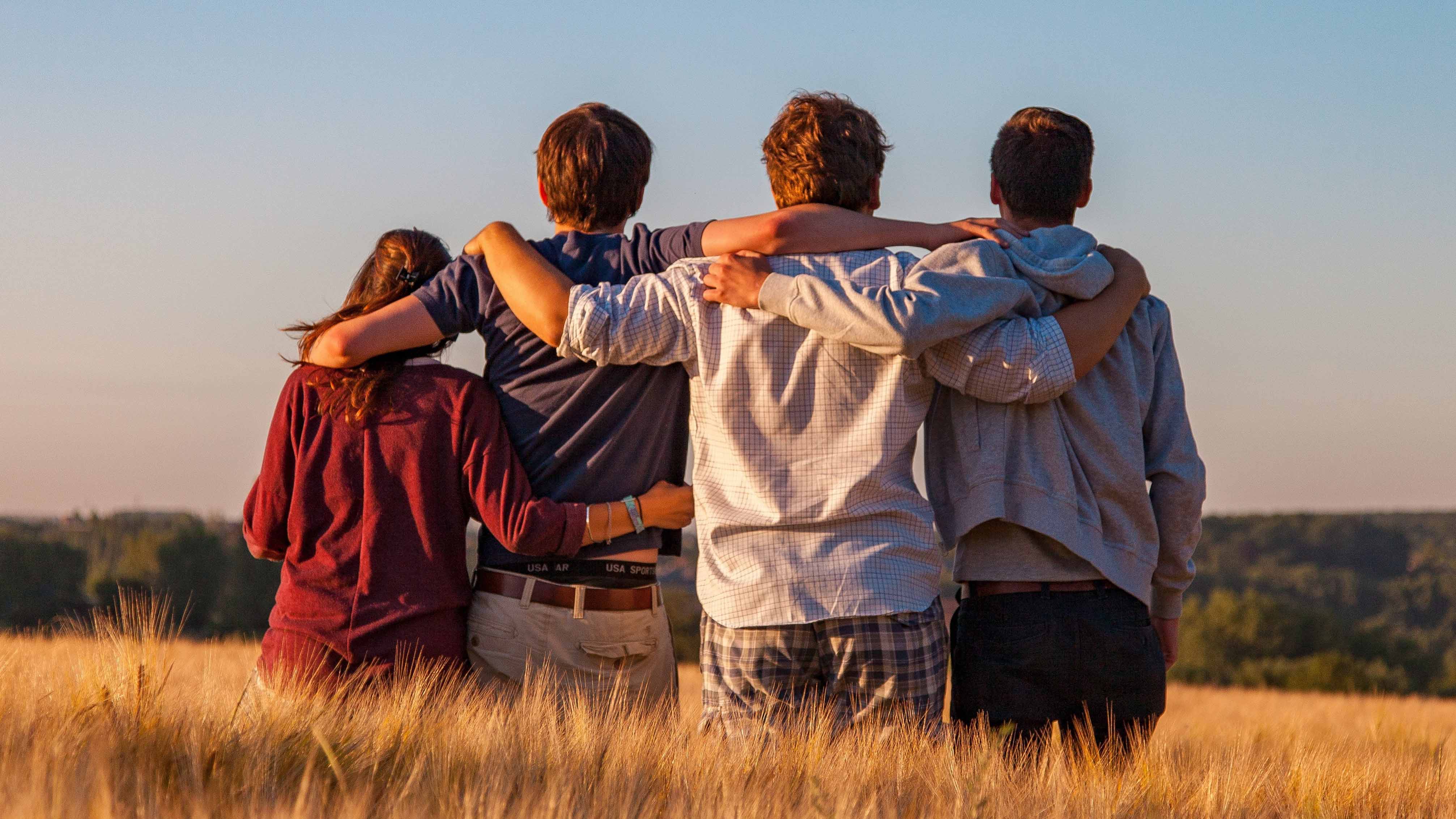 Vier junge Menschen stehen eng umschlungen mit dem Rücken zur Kamera in einem goldgelben Kornfeld. Sie haben sich gegenseitig die Arme um die Schultern gelegt und blicken im warmen Licht der Abendsonne gemeinsam in die Ferne.