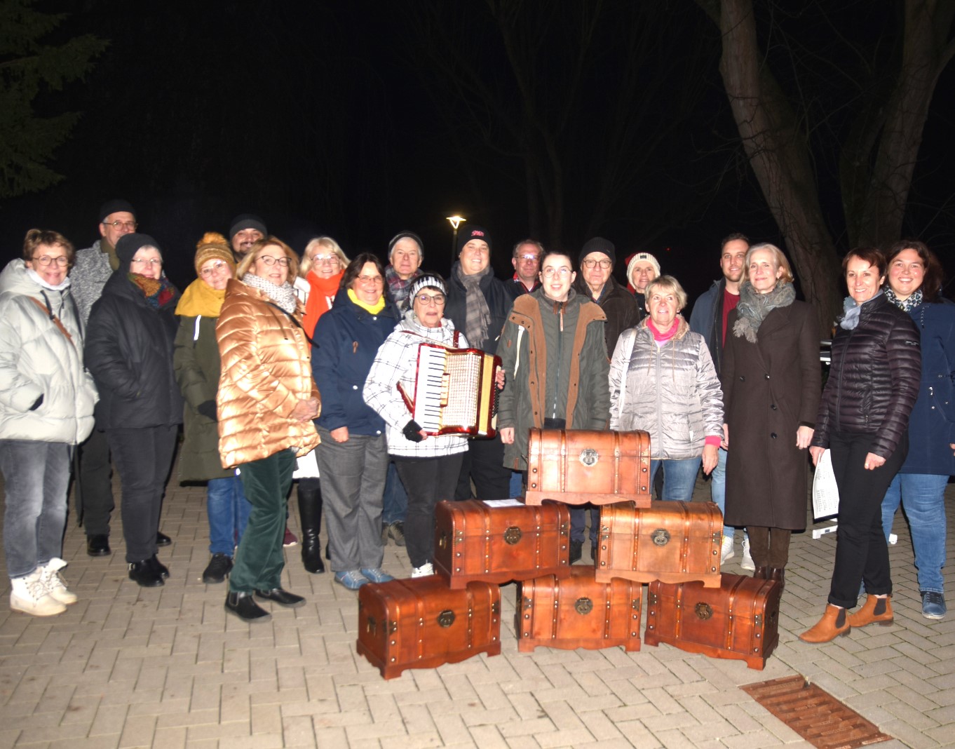 Eine Gruppe Menschen, darunter Karola Zühlke mit Akkordeon, steht im dunklen Hof des Margaretha-Pistorius-Hauses, vor sich sechs Schatzkisten.