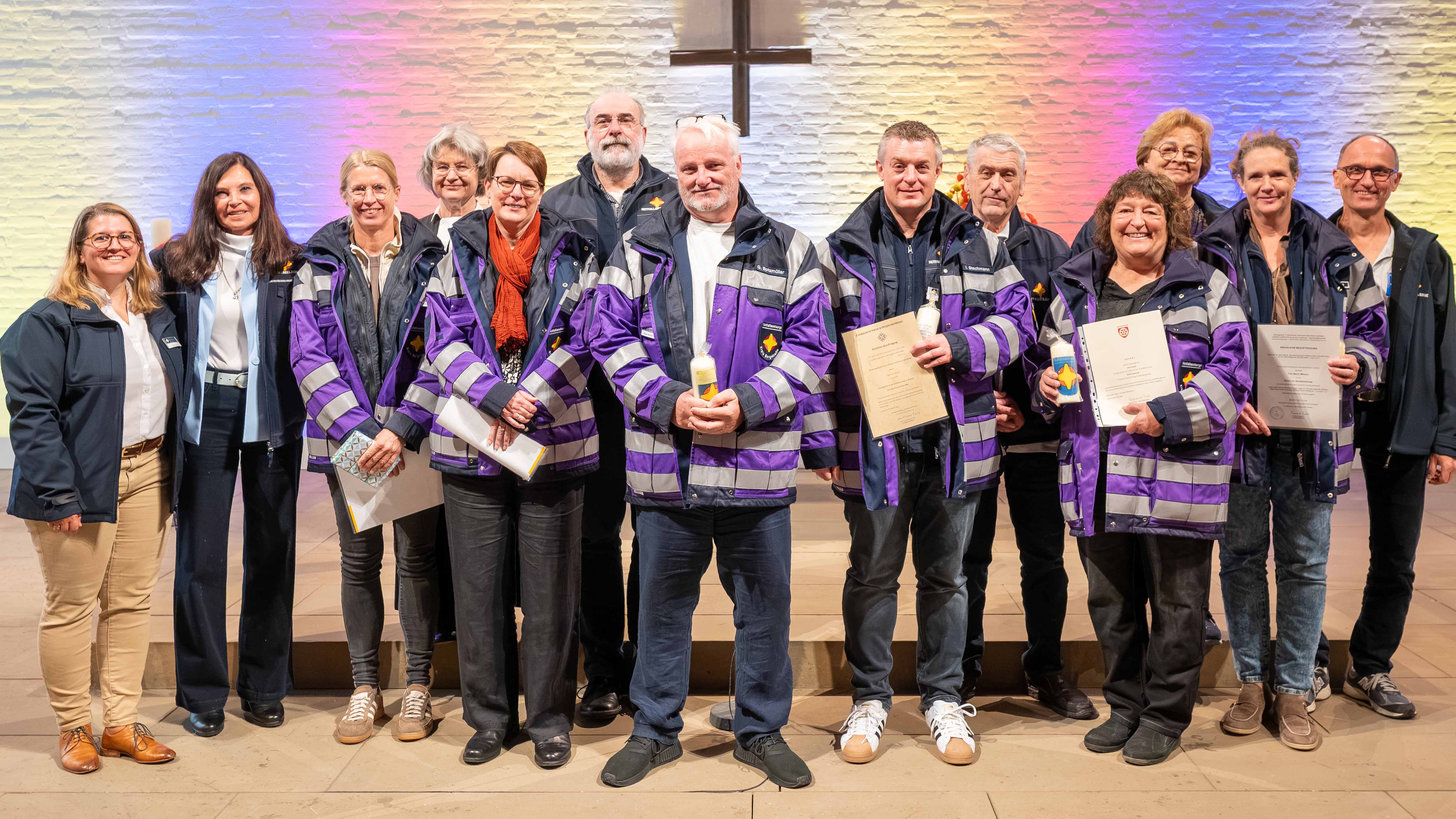 Gruppenfoto von 13 Frauen und Männern im Altarraum einer Kirche. Mehrere Personen tragen violett-dunkelblaue Einsatzjacken mit reflektierenden Streifen der Notfallseelsorge. Einige halten Urkunden oder verzierte Kerzen in den Händen. Die Gruppe steht in zwei Reihen vor einer Natursteinwand, die in Rosa, Blau und warmen Gelbtönen beleuchtet ist. Über den Köpfen der Personen hängt ein großes Holzkreuz.