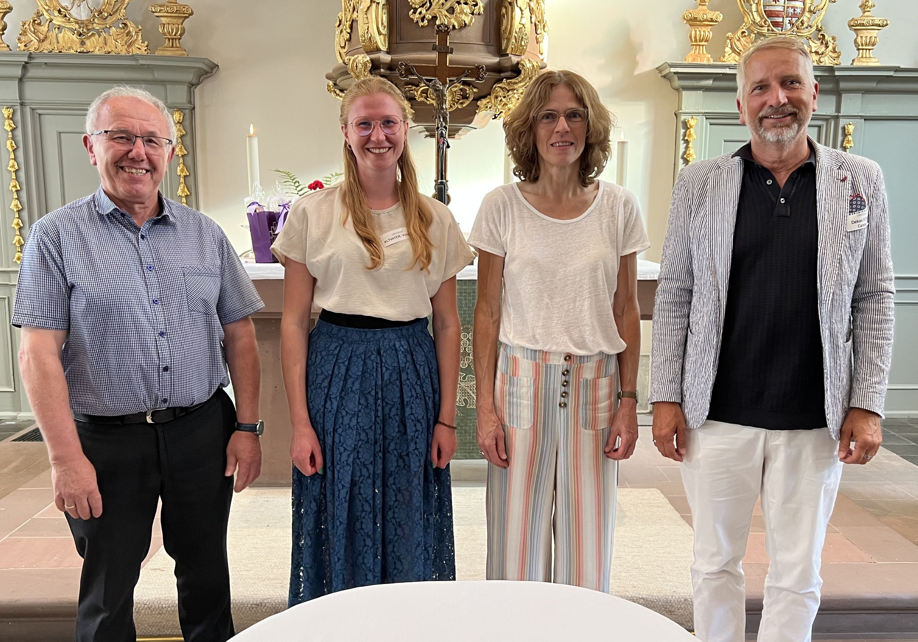 Vier Menschen stehen vor einem Altar in einer barock anmutenden Kirche. Sie schauen allesamt freundlich drein.