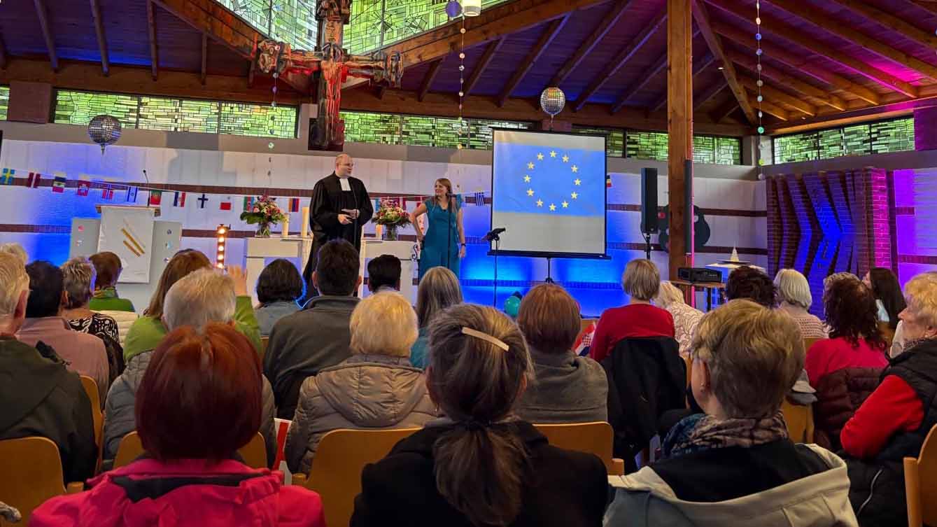 Menschen sitzen in einer Kirche und verfolgen eine Veranstaltung auf einer Bühne mit zwei sprechenden Personen und einer projizierten EU-Flagge.