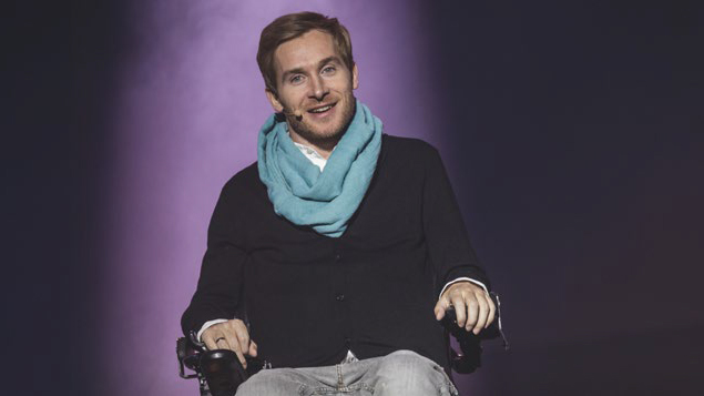Koch_Samuel_C_Promikon_16zu9.jpg Eine Person sitzt mittig auf einer Bühne vor dunklem Hintergrund mit violettem Licht. Sie trägt ein dunkles Oberteil, helle Hose und einen auffälligen hellblauen Schal. An der Wange ist ein Headset-Mikrofon befestigt, die Person blickt lächelnd ins Publikum.