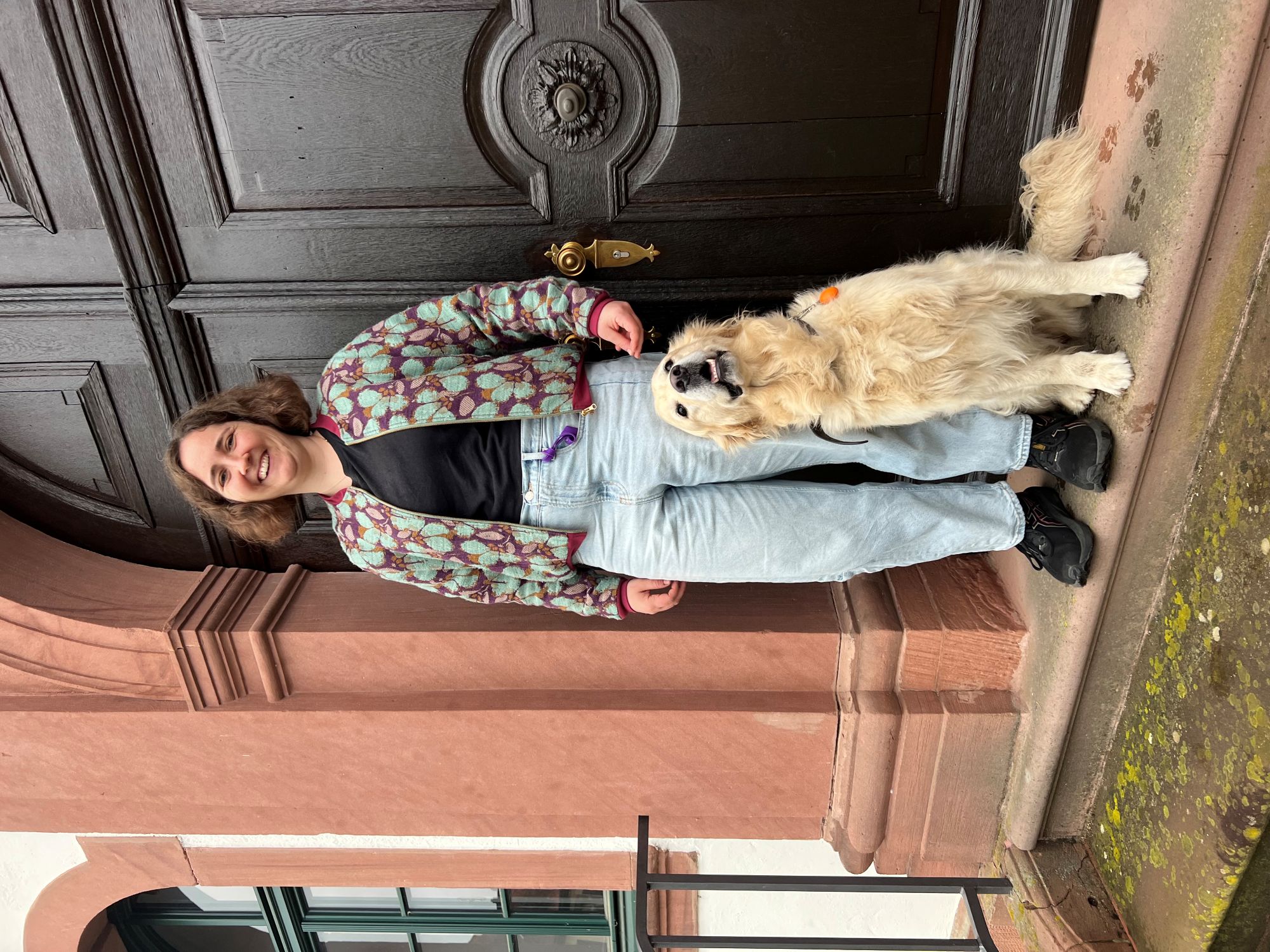 Eine Frau steht vor einer historischen Tür und schaut freundlich drein. Vor ihr sitzt ein Hund der Rasse Golden Retriever.