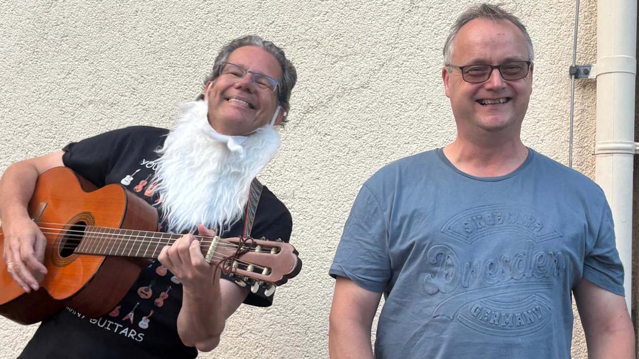 Zwei lachende Männer stehen vor einer Hauswand; einer trägt einen weißen Kunstbart und spielt Gitarre, der andere steht daneben.