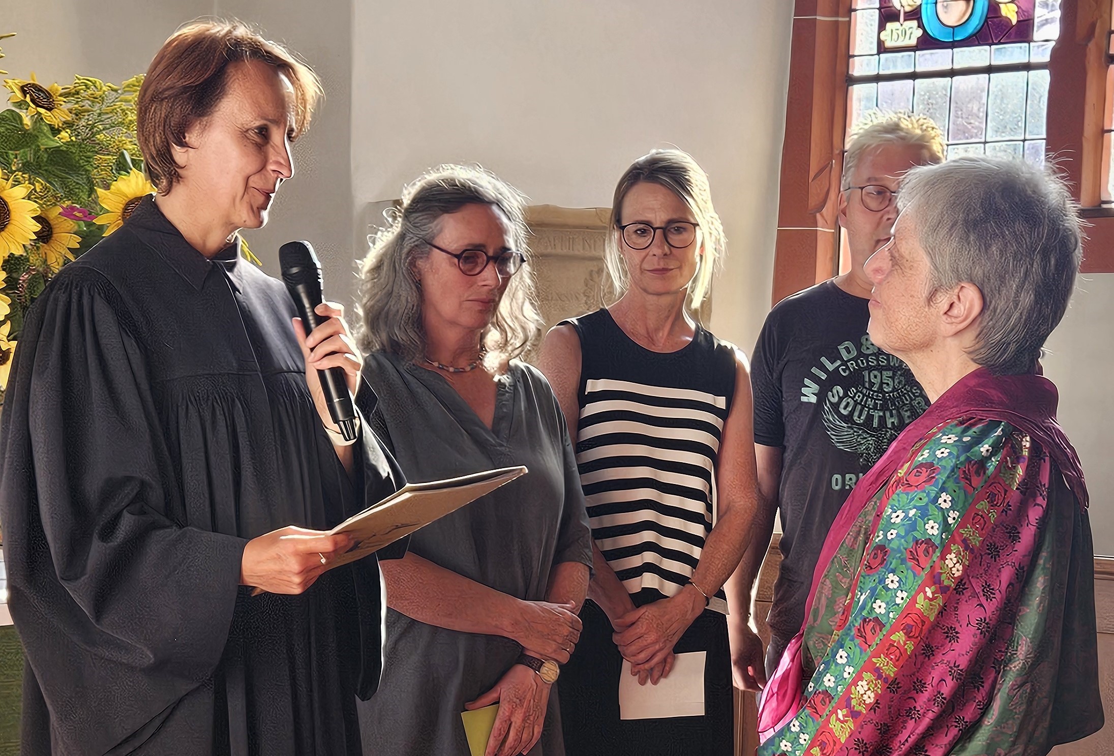 Dekanin Birgit Hamrich im Talar und Renate Nagel-Kroll in einem bunten Kleid, stehen sich am Altar der Niddaer Stadtkirche gegenüber. Dekanin Hamrich hält ein Mikrofon und eine Mappe in den Händen. Im Hintergrund stehen Kolleginnen aus dem Gemeindepädagogischen Dienst.