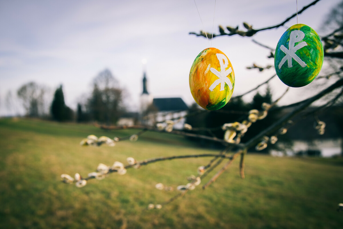 Bunt bemalte Ostereier hängen an einem Baum. Im Hintergrund ist eine Dorfkirche zu sehen.