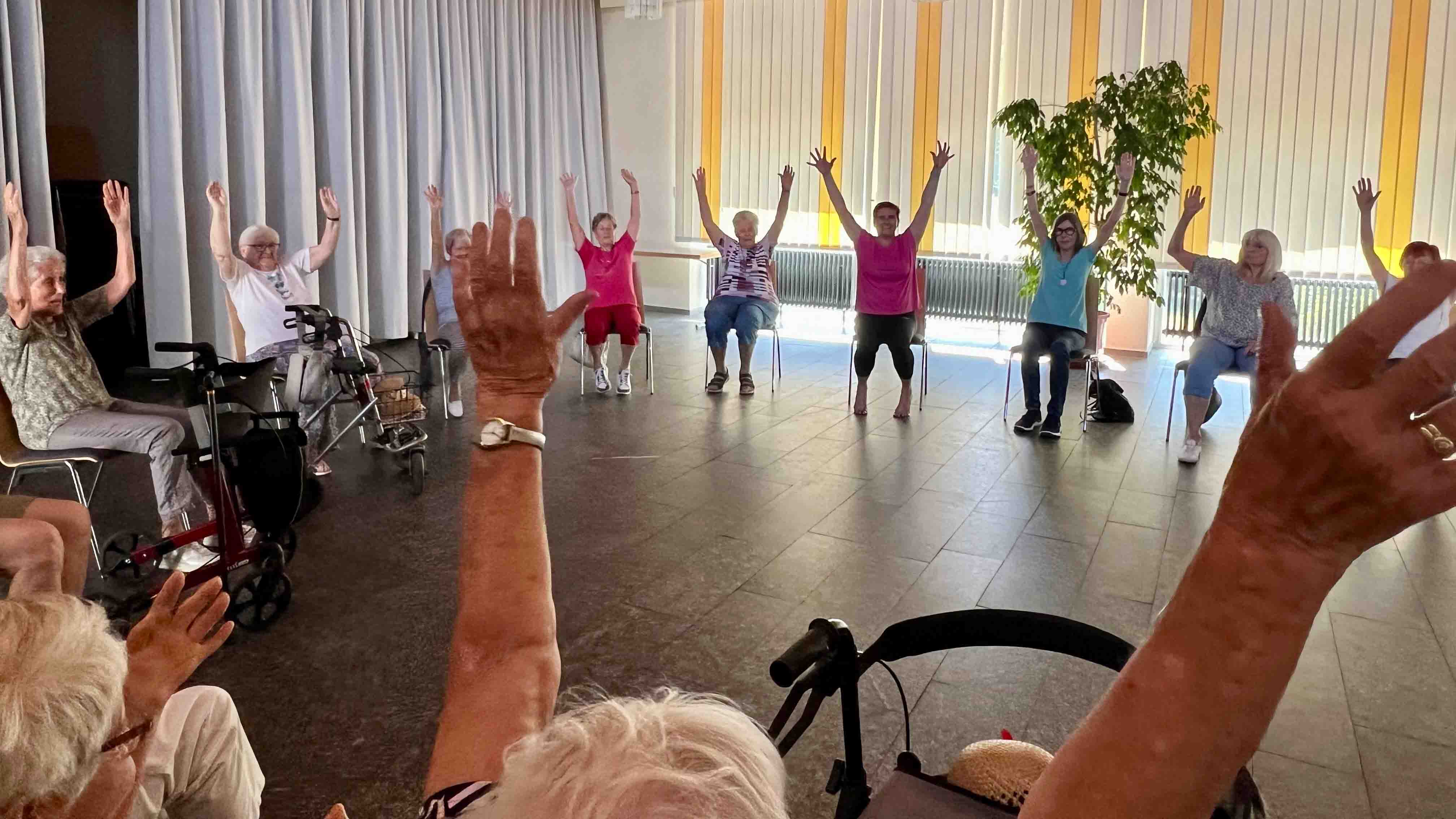 bzw_Bewegungstreffpunkt_C_Evang_Kirchengemeinde_Zwingenberg_2_16zu9.jpg Ältere Menschen sitzen im Kreis und heben bei einer gemeinsamen Bewegungsübung die Arme, einige nutzen einen Rollator oder sitzen auf einem Stuhl.